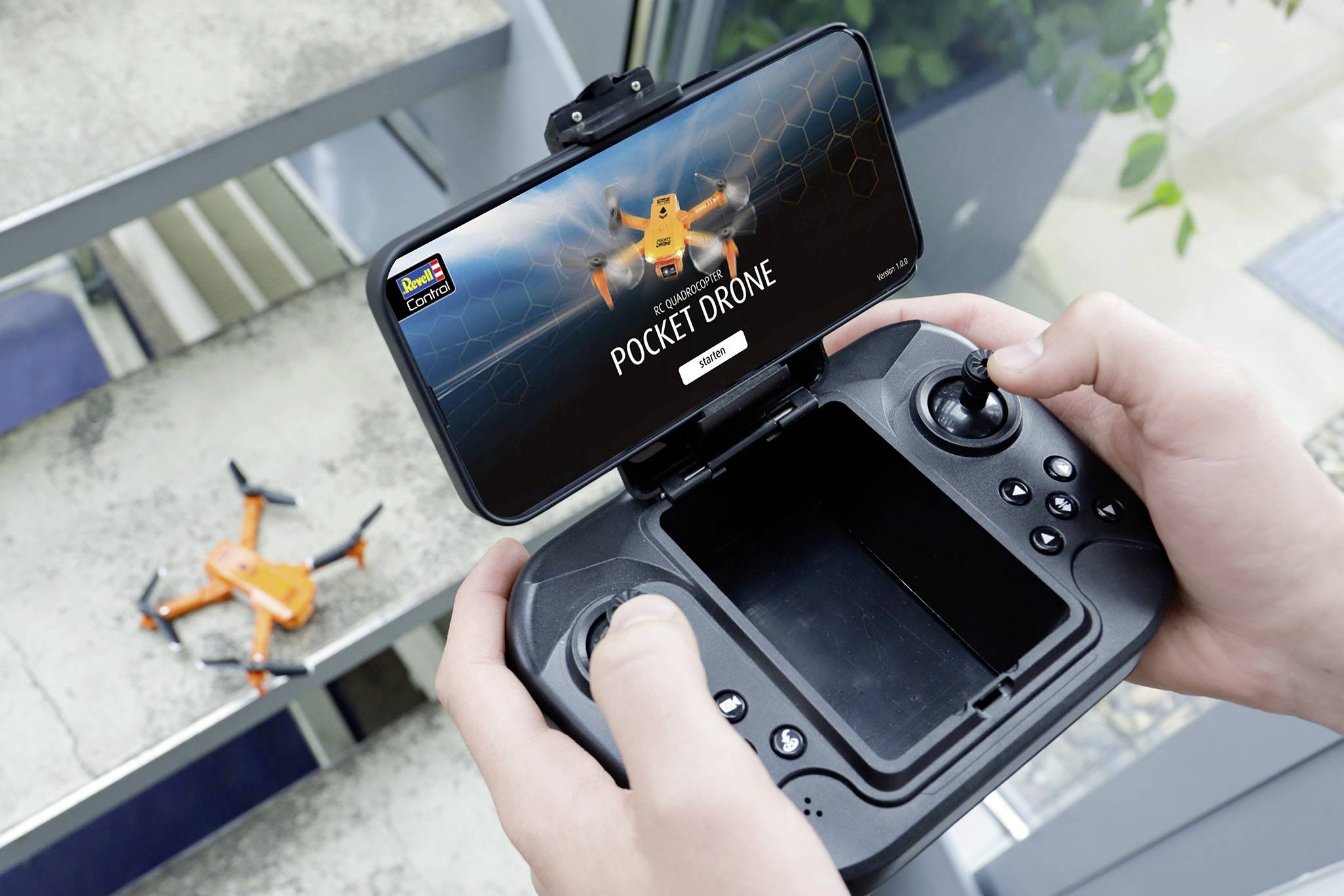 Oseba drži daljinski upravljalnik za brezpilotni letalnik z zaslonom pametnega telefona, na katerem piše 'Pocket Drone'. V ozadju je majhna oranžna brezpilotna letalo na stopnicah.