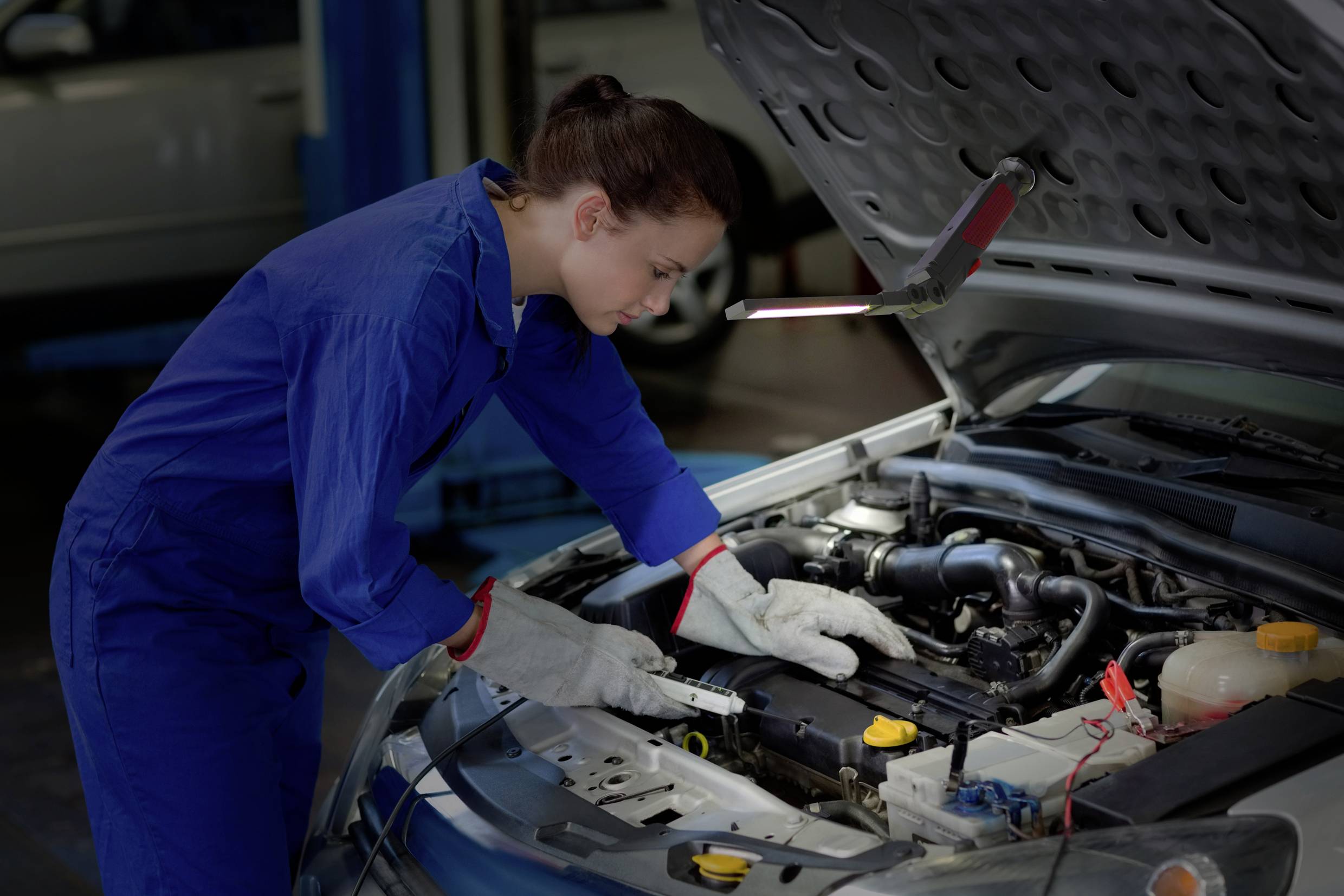 Ženska v modri delovni obleki popravlja pod motornim pokrovom avtomobila v delavnici.