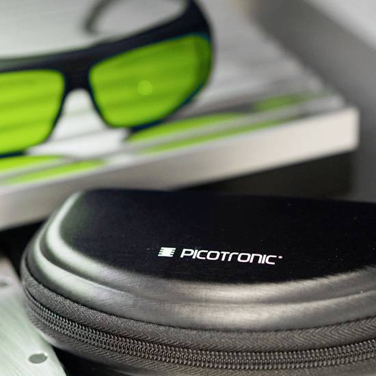 Etui z napisom 'PCTRONIC' in zelena zaščitna očala, ki ležita na mizi.