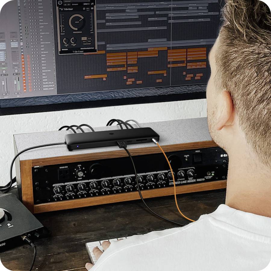 Oseba dela na računalniku z velikim monitorjem, ki prikazuje programsko opremo za glasbeno produkcijo. V ospredju so audio naprave.