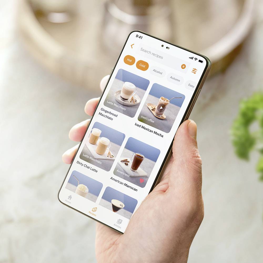 Ena roka drži pametni telefon, ki prikazuje izbor pijač v aplikaciji, med katerimi so café latte, kava in mlečni koktajli.