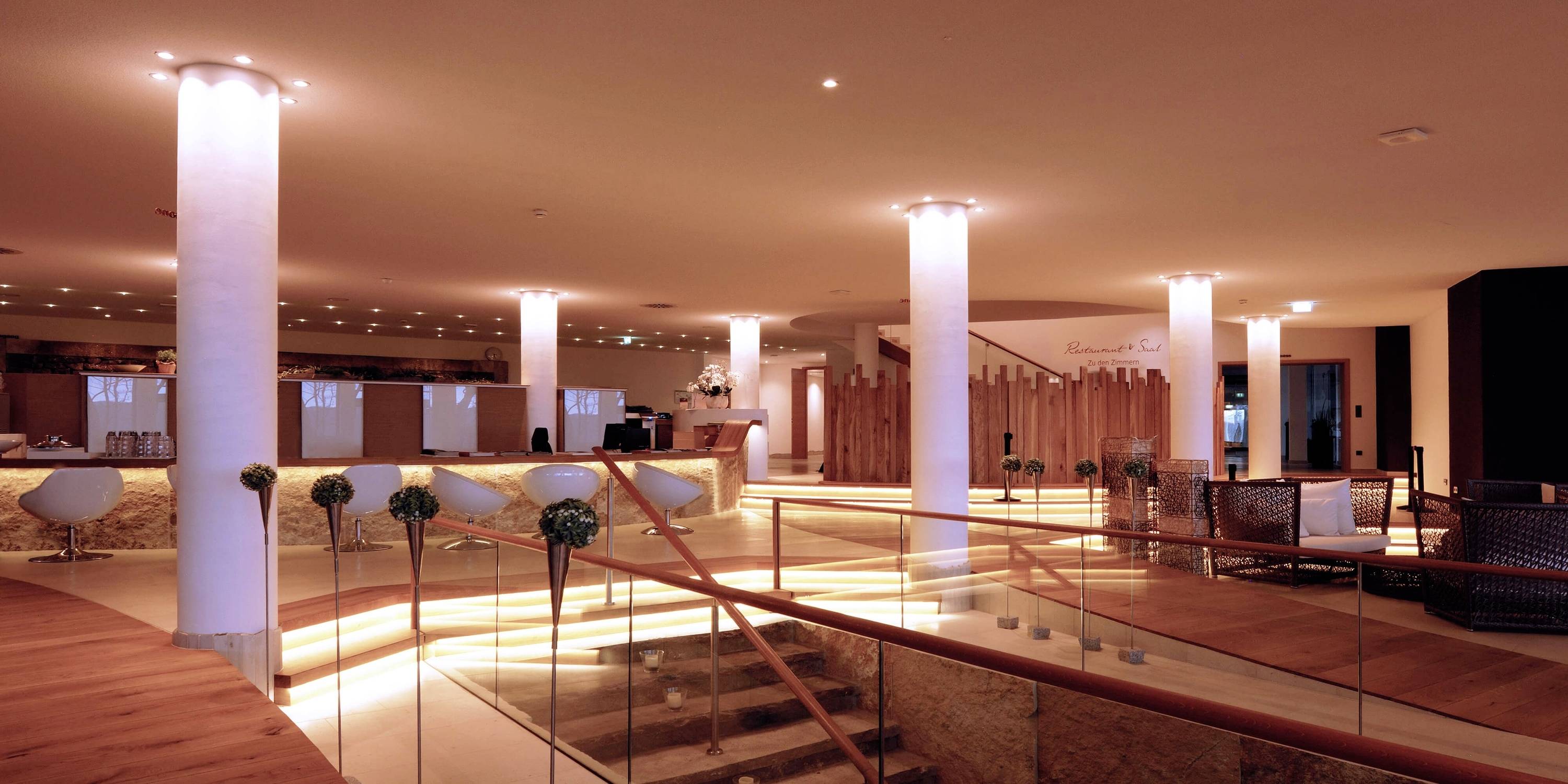 Moderna hotelska lobby z toplo osvetlitvijo, vijugastimi stopnicami, rdečimi stebri in več sedeži. V ozadju je recepcija.