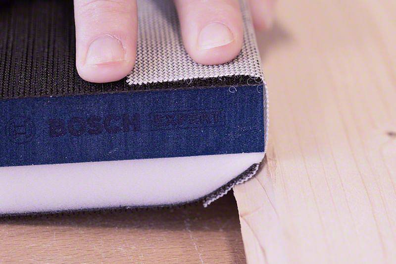 Ena roka drži brusilni stroj z brusilnim trakom na kosu lesa. Na napravi je vidna znamka 'Bosch Expert'.
