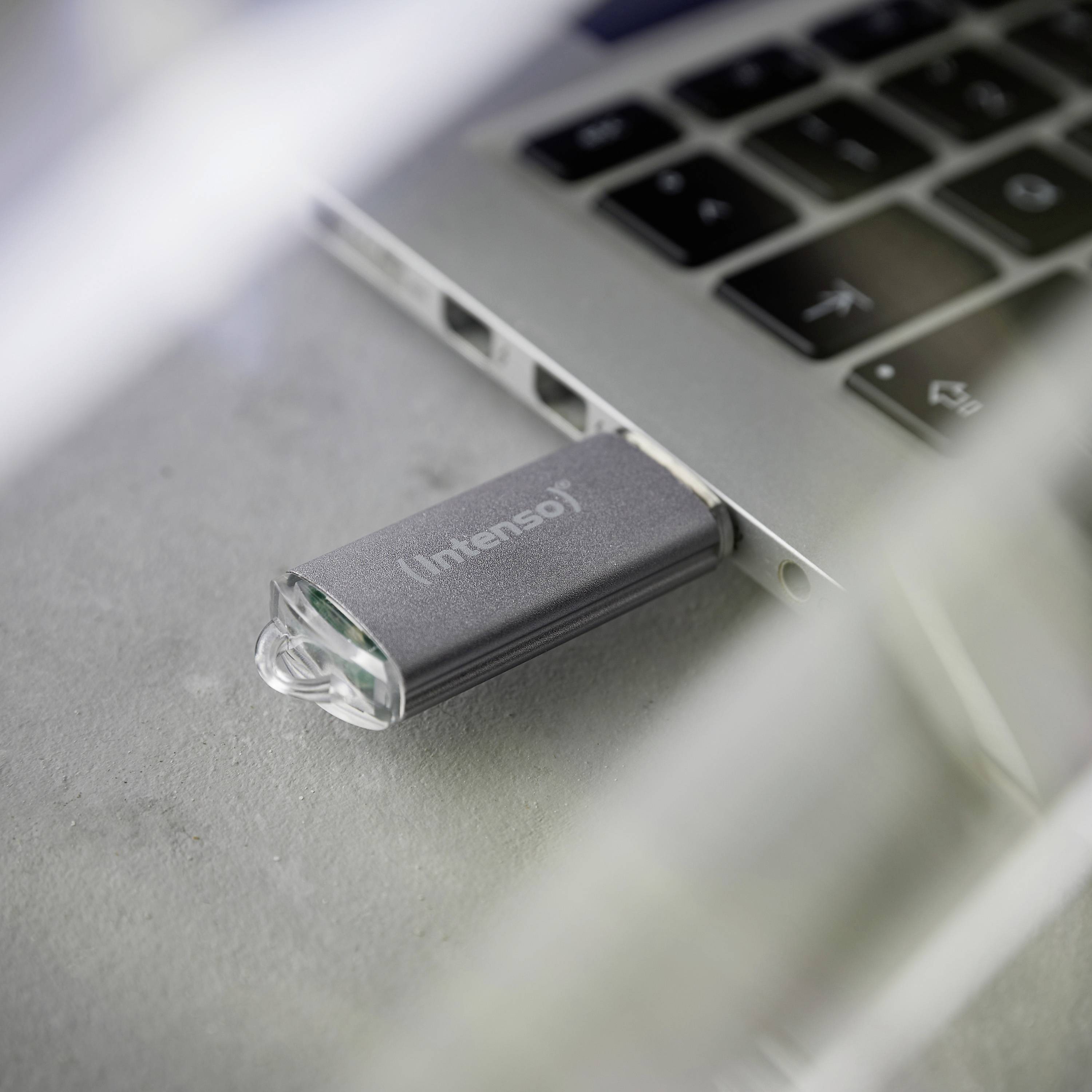 USB ključek je priklopljen na prenosni računalnik. Ključek leži na sivi podlagi in ima napis 'Intenso'.