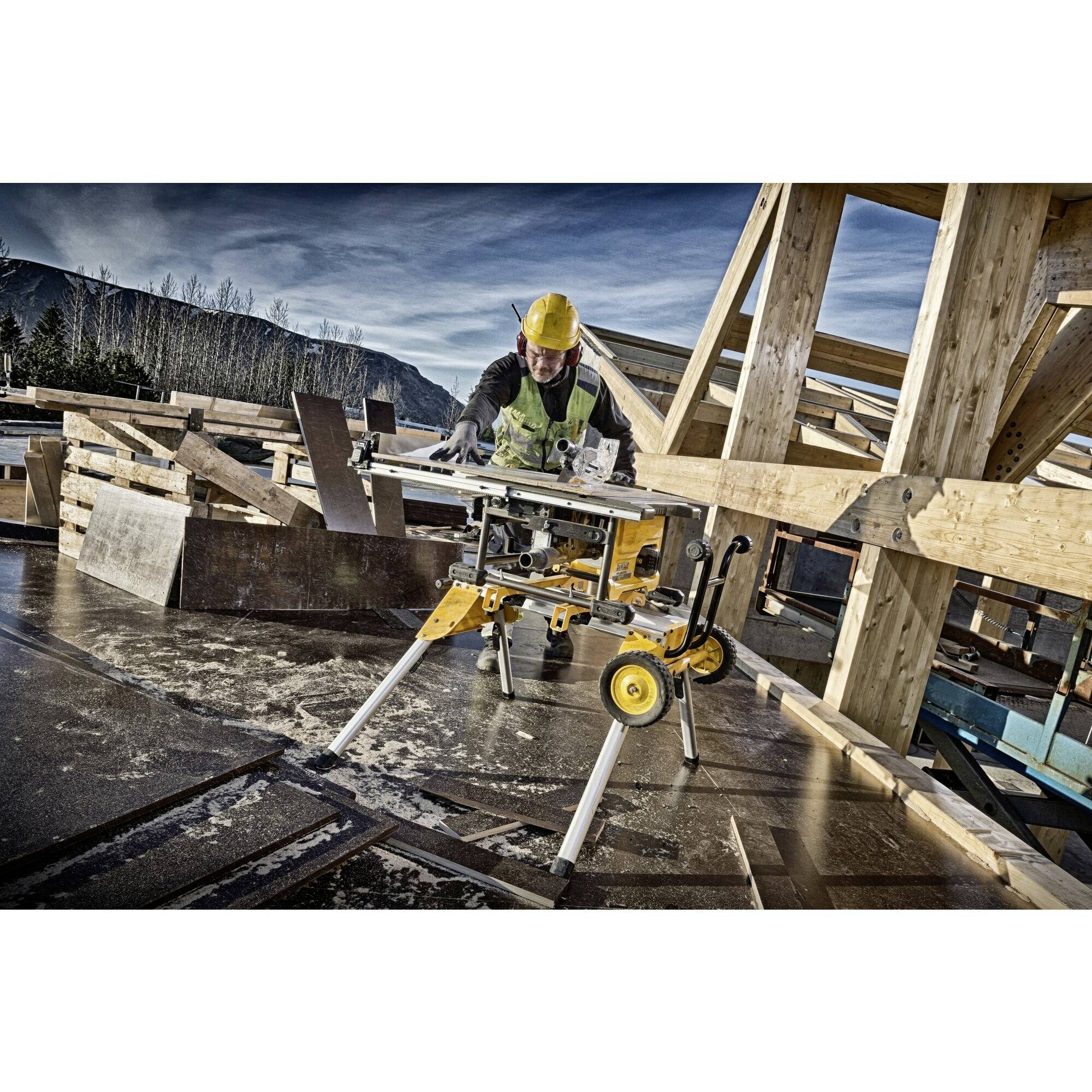 Delavec z rumeno čelado in zaščitnim jopičem upravlja krožno žago na gradbišču. Lesena konstrukcija v ozadju, jasen dan.