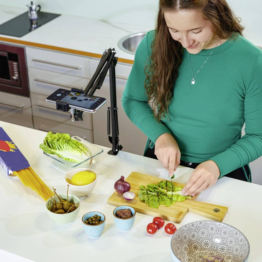 Oseba reže sveže zelenjavo v moderni kuhinji, obkrožena z ingrediencami, kot so limone, peteršilj, čebula in paradižniki.