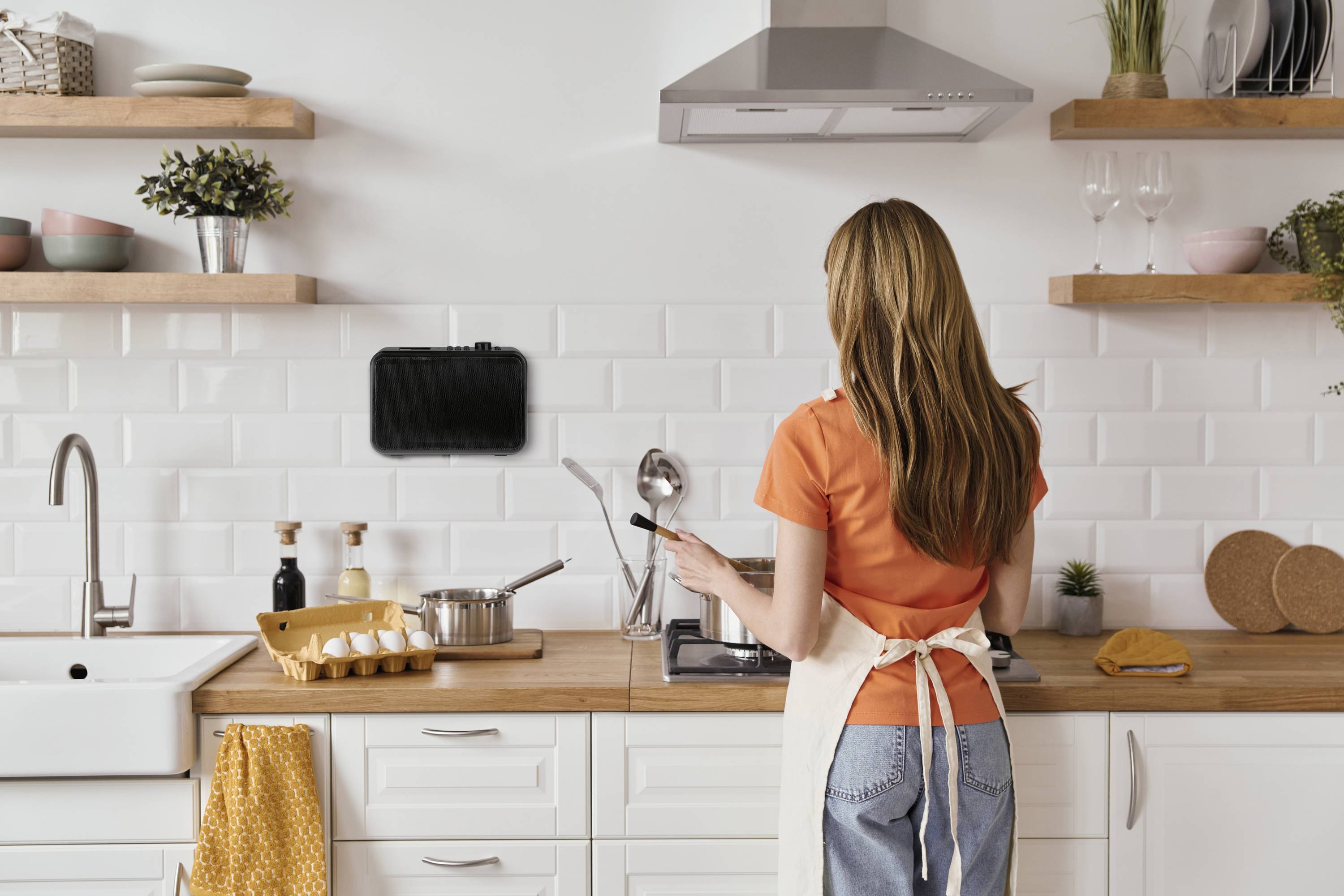Ženska v oranžni majici in predpasniku kuha v moderni kuhinji. Uporablja tablico kot navodila za recept, pritrjeno na steno.
