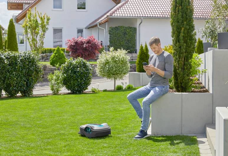 Moški sedi na vrtu na zidu in s pametnim telefonom upravlja robotsko kosilnico, ki deluje na skrbno pokošeni tratni površini v stanovanjski soseski.