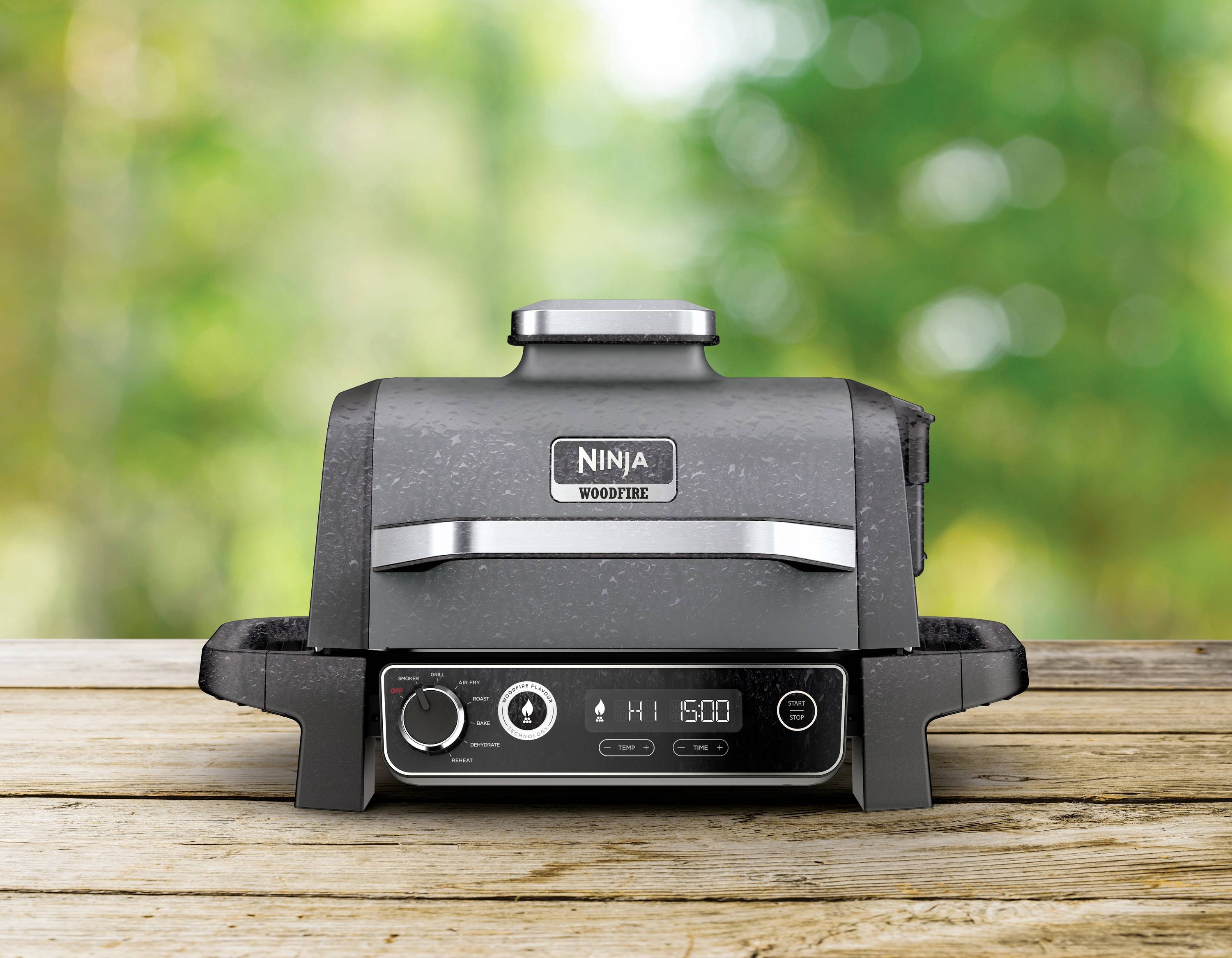 Kompakten črn žar znamke 'Ninja Woodfire' na leseni mizi pred zamegljeno zeleno ozadje.