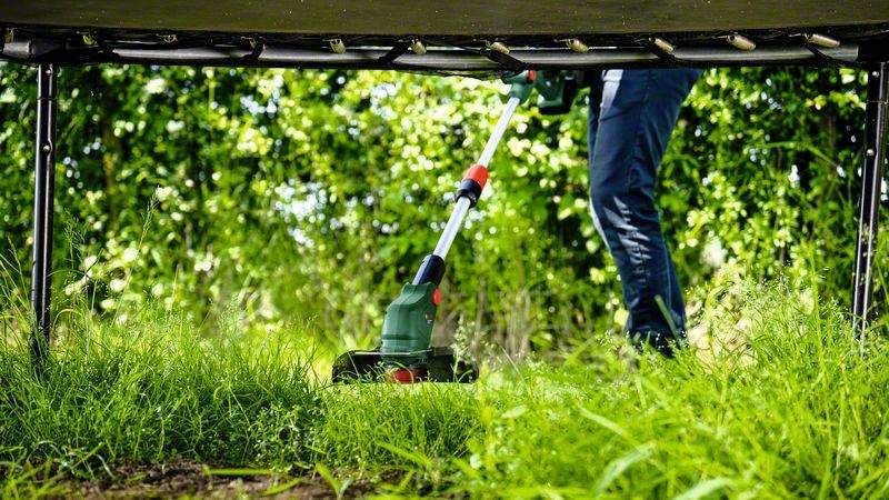 Oseba s električnim trimmerjem kosi visoko travo pod trampolinom. V ozadju so vidni zeleni grmi.