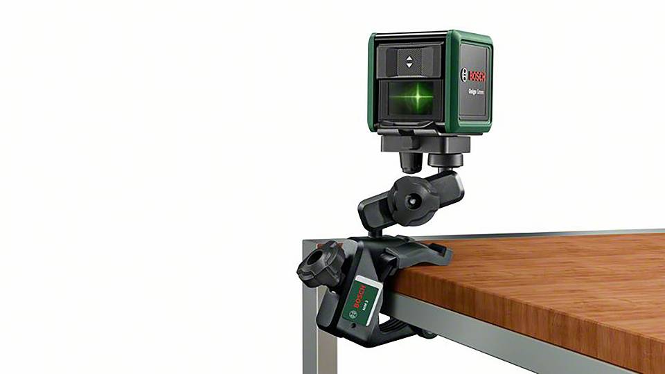 Križni linijski laser na stojalu, pritrjen na rob mize. Naprava projicira zeleno križno linijo za natančno poravnavo.