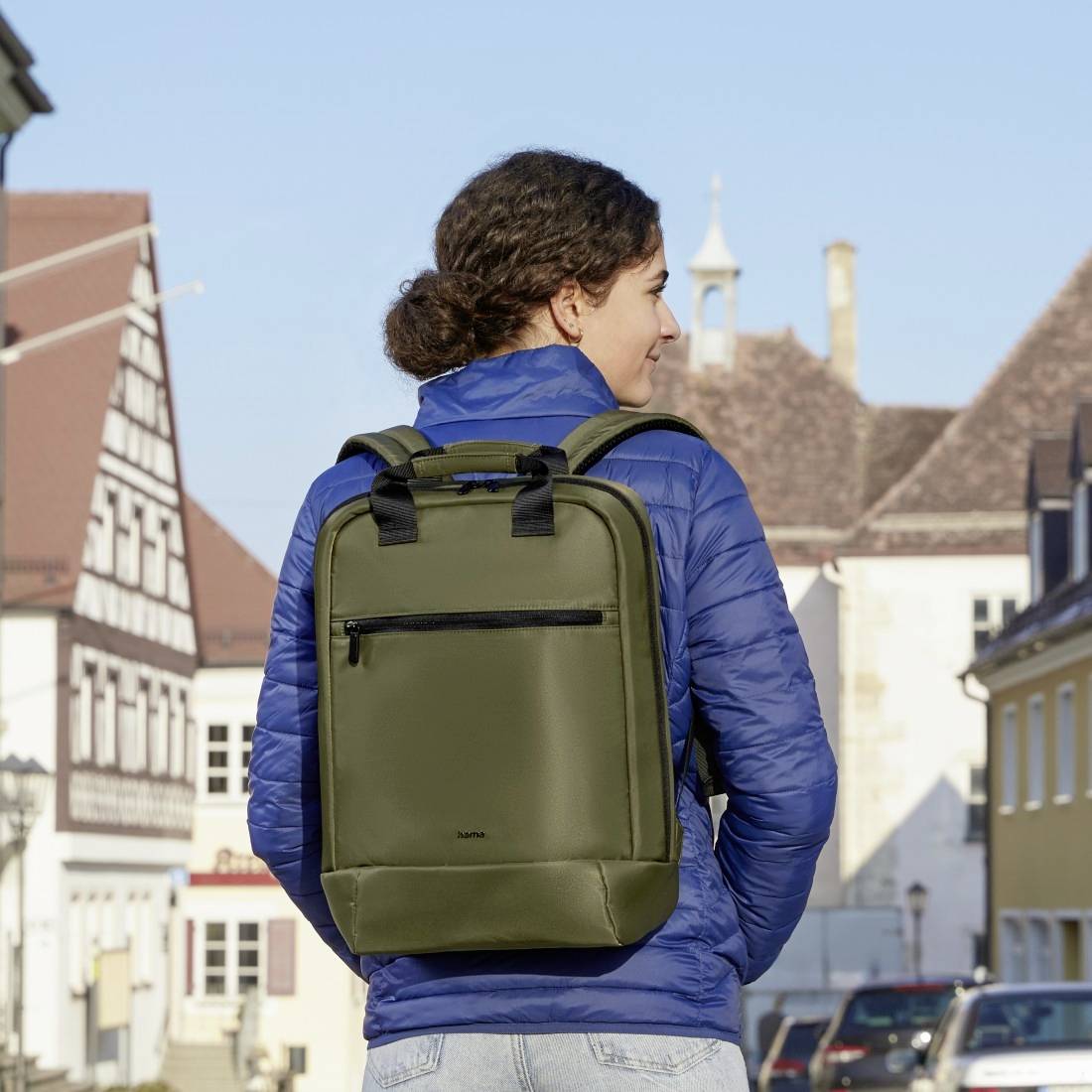 Človek v modrem anoraku nosi olivno zelen nahrbtnik in stoji pred zgodovinskimi stavbami v urbanem okolju.