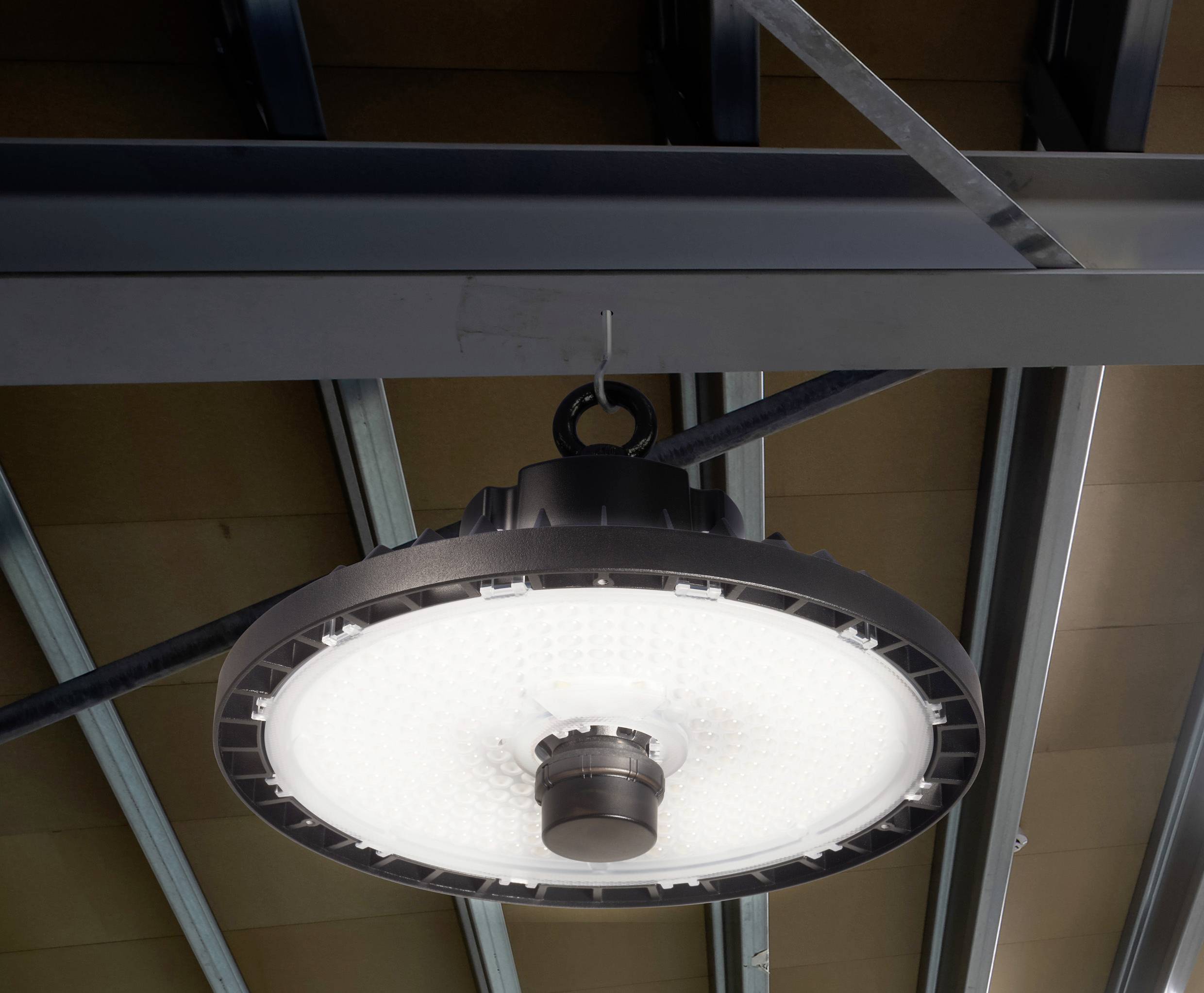 Stropna svetilka z velikim, okroglim senčnikom in LED-osvetlitvijo, pritrjena na kovinski konstrukciji pod stropom dvorane.