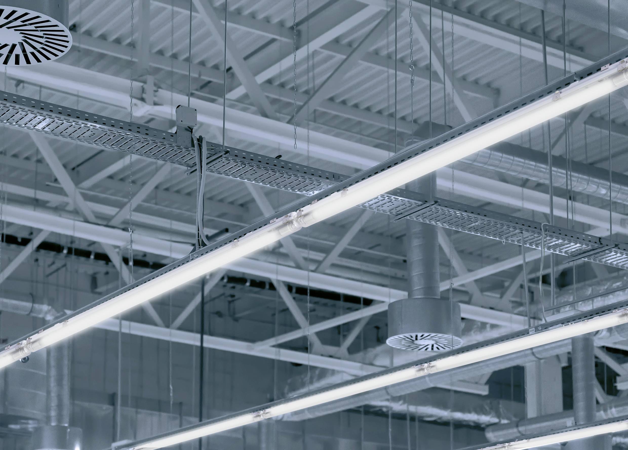 Svetle LED fluorescentne cevi na industrijski stropu z vidnimi prezračevalnimi kanali in kabli.