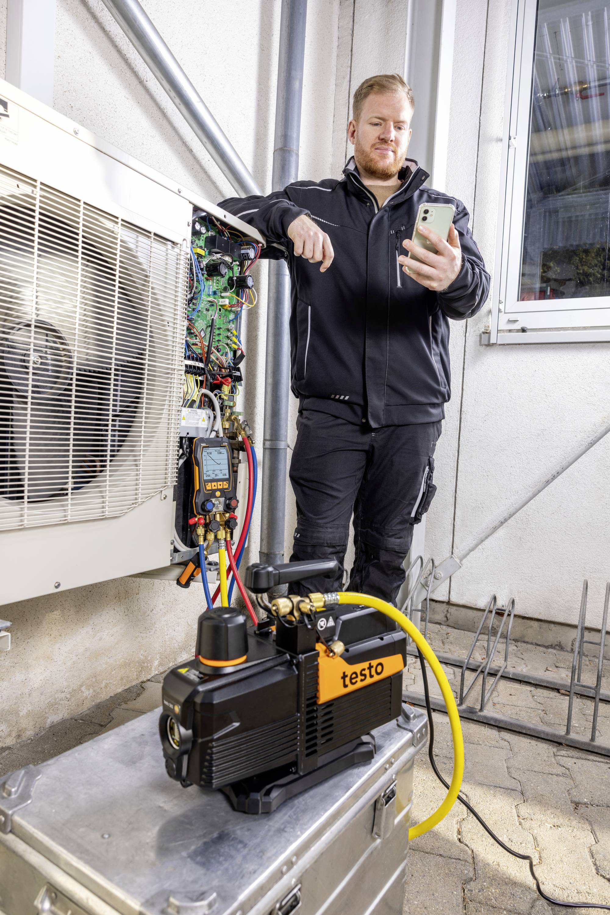 Tehnik stoji poleg klimatske naprave in v roki drži pametni telefon. Na klimatski napravi je priključena merilna naprava, pred njo pa leži vakuumska črpalka.