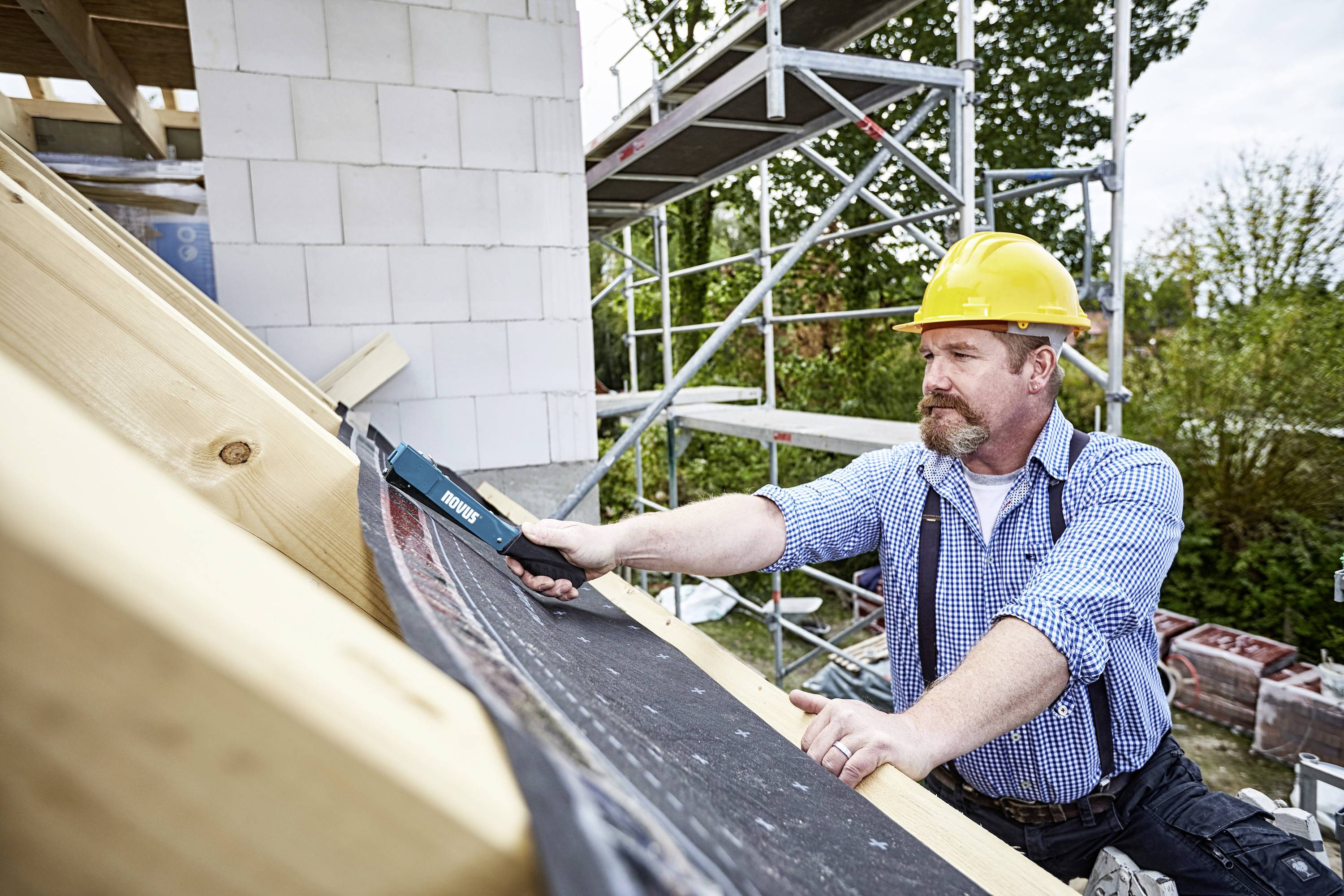 Delavec z rumeno zaščitno čelado pritrjuje streho desko. V ozadju so odri in stavba v gradnji.