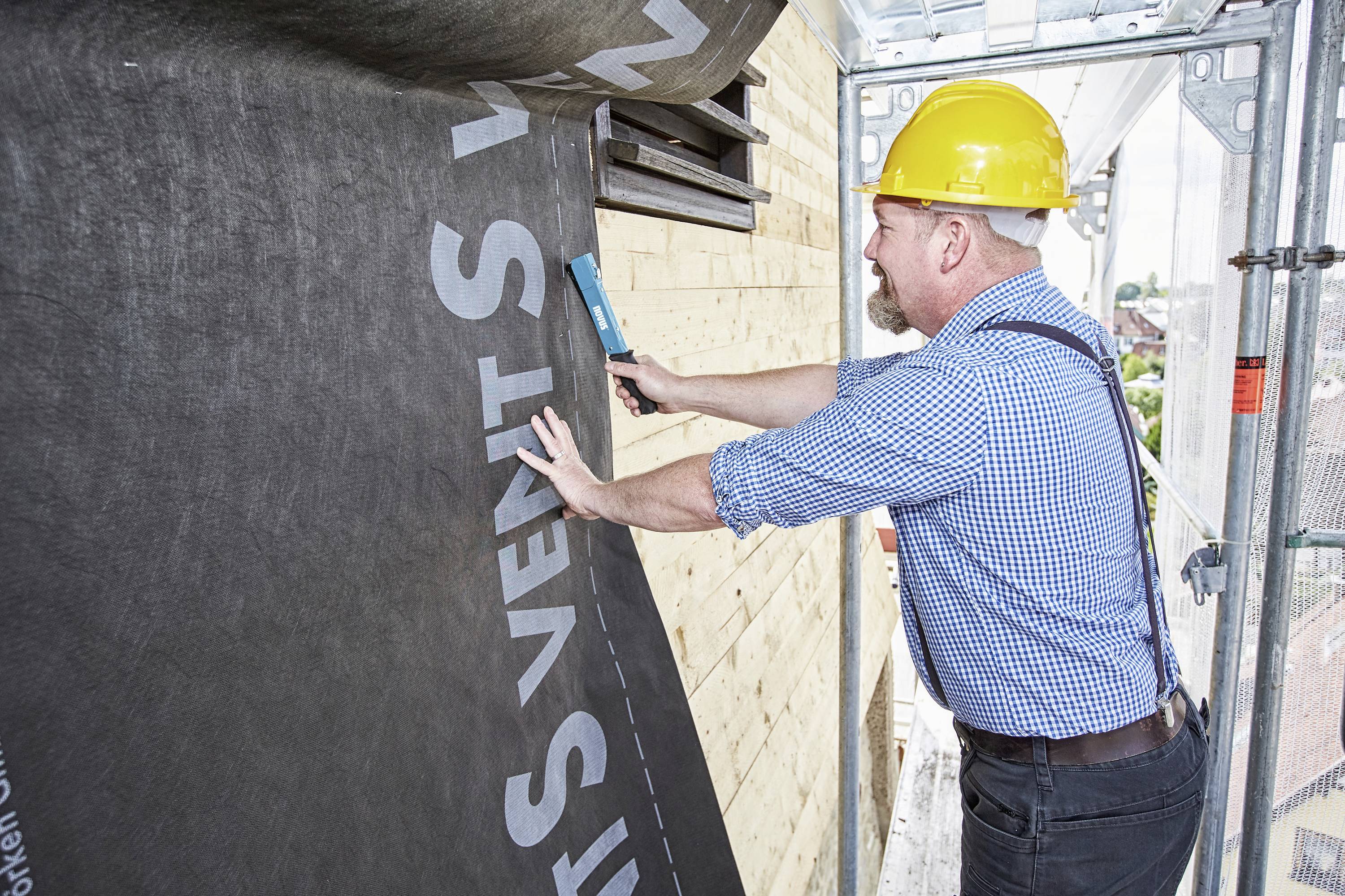 Delavec v gradbeništvu nosi zaščitno čelado in pritrjuje parna zaporo na zunanji steni stavbe na gradbišču.