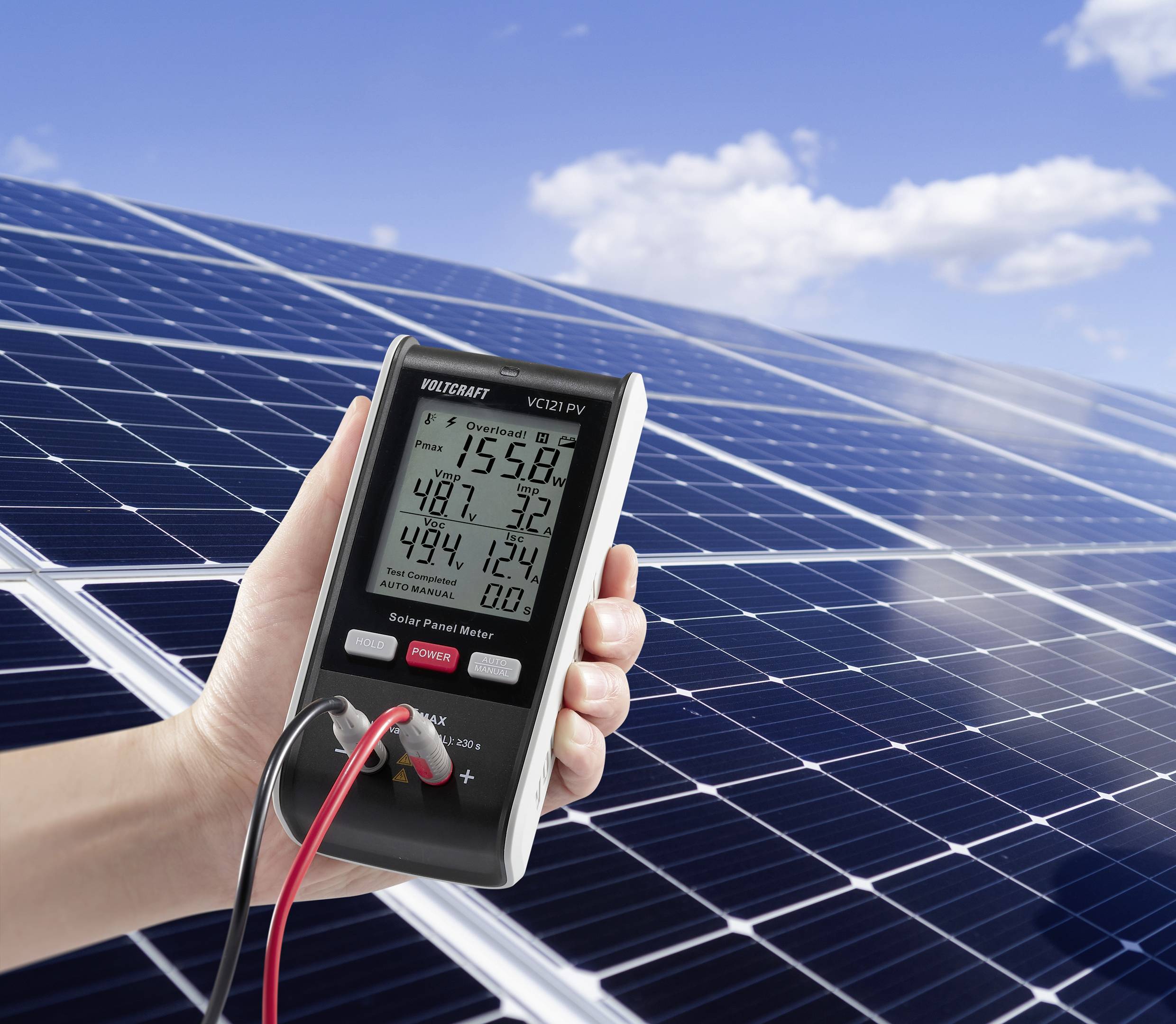 Roka drži merilnik solarnih panelov pred velikim solarnim panelom; naprava prikazuje električne meritve.