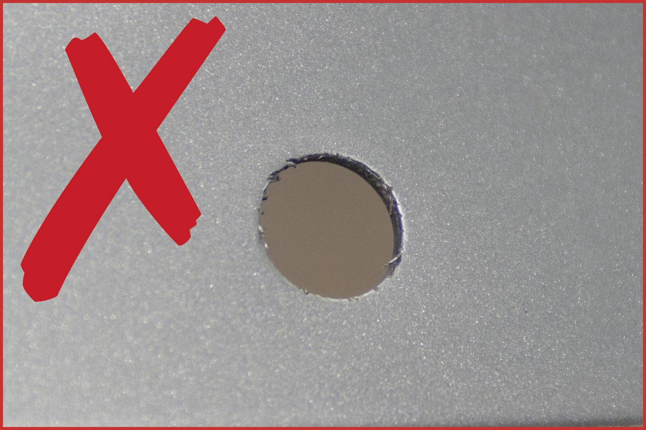 Rdeč 'X' poleg luknje na sivi površini, verjetno za označitev, da je luknja nezaželena ali napačna.