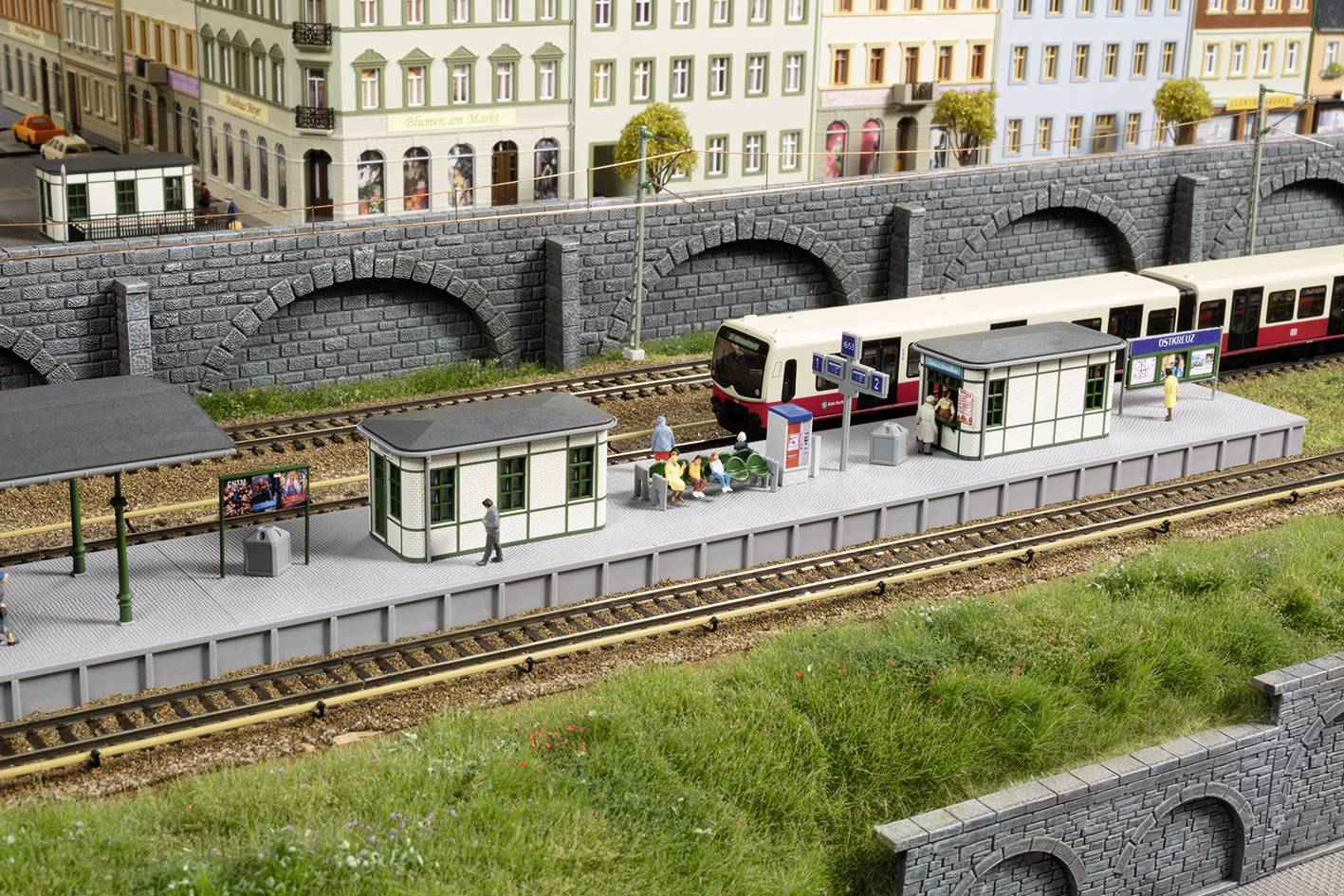Majhna scena modelne železnice z mestno postajo, peronom, miniaturnimi vlaki in ljudmi. Stavbe v ozadju.