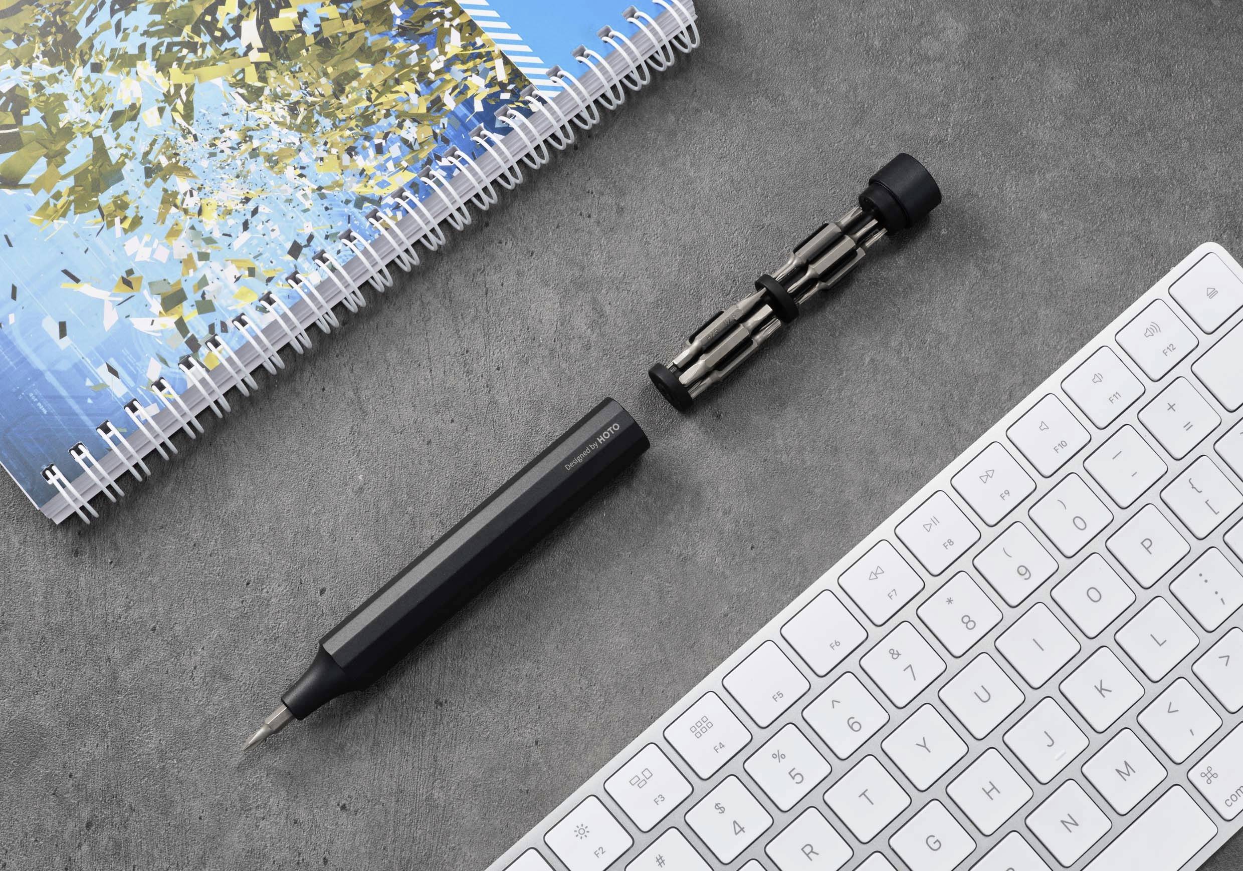 Beležnica z modrim ovitkom, izvijač z nastavkom in računalniška tipkovnica na sivi podlagi.