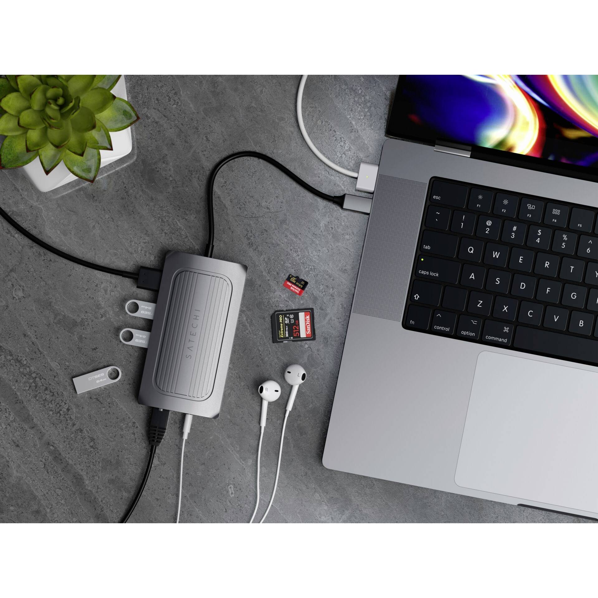 Prenosni računalnik s priključenim USB razdelilcem, pomnilniškimi karticami, slušalkami in USB pogoni na pisalni mizi z rastlino.