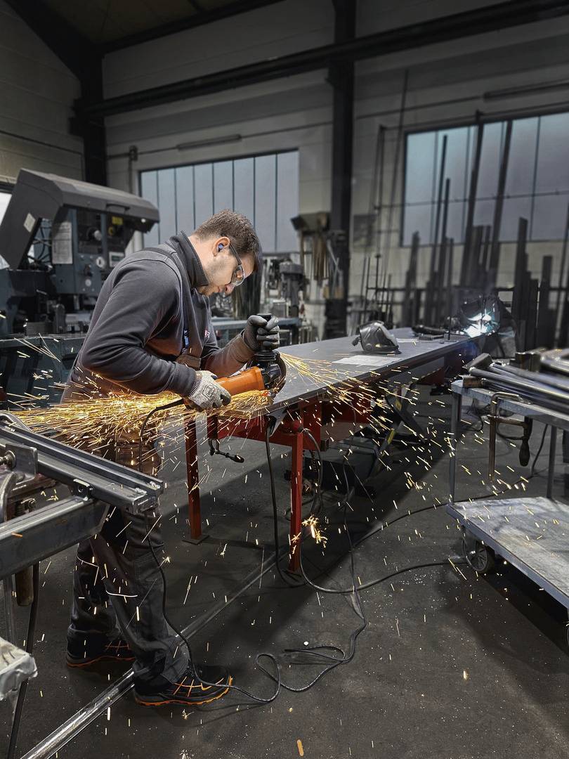 Delavec nosi zaščito za sluh in brusi kovino v delavnici. Z brusilnega stroja letijo iskre. V ozadju so vidni industrijski stroji.