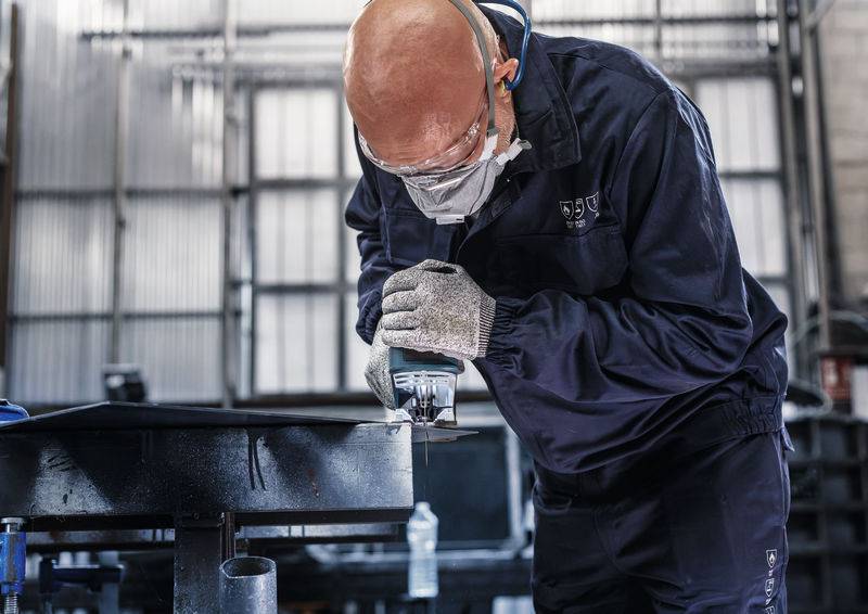 Oseba dela z varnostno opremo v delavnici, nagiba se čez mizo in uporablja orodje. Industrijsko ozračje.