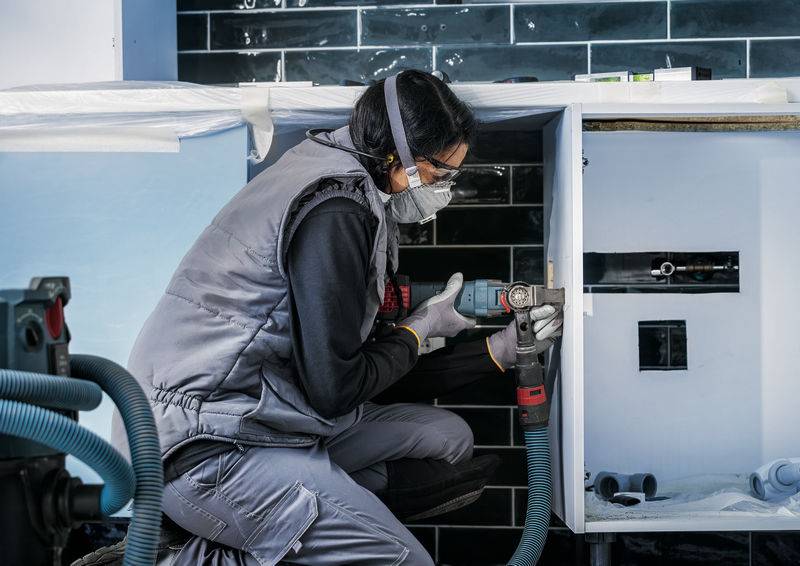 Oseba v delovni obleki popravlja vrata omare v delavnici. Nosi zaščitna očala in masko ter uporablja električno orodje.