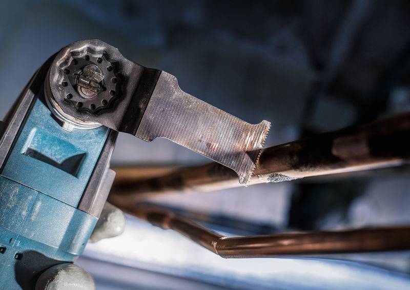 Stroj reže bakrene cevi v industrijskem okolju. Cevi potekajo horizontalno, stroj pa ima kovinsko žago.