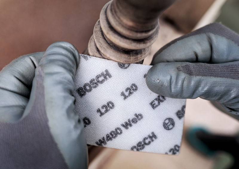 Oseba v sivih rokavicah brusi košček lesa z brusilnim papirjem znamke Bosch.
