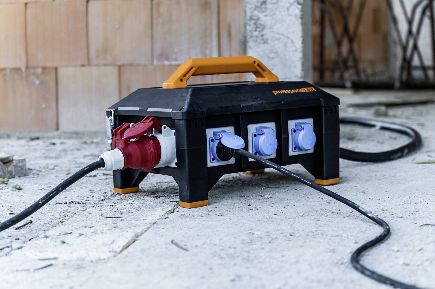 Mobilna razdelilna električna omarica za gradbišče z več modrimi vtičnicami in rdečim industrijskim vtikačem. Stoji na betonskih tleh.