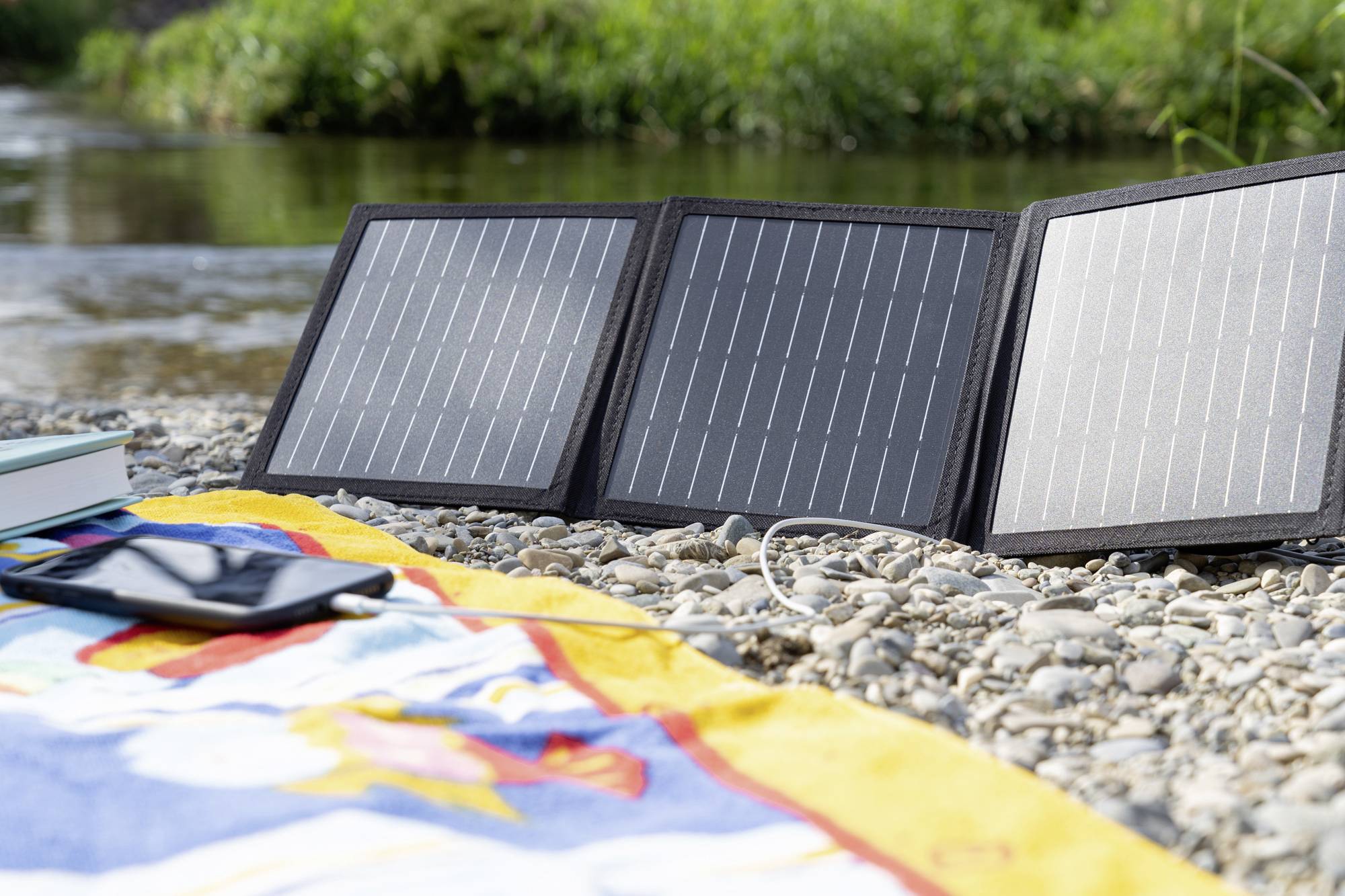 Prenosni solarni panel leži na produ ob rečnem bregu. Zraven je pisano brisačo in zaprta knjiga.