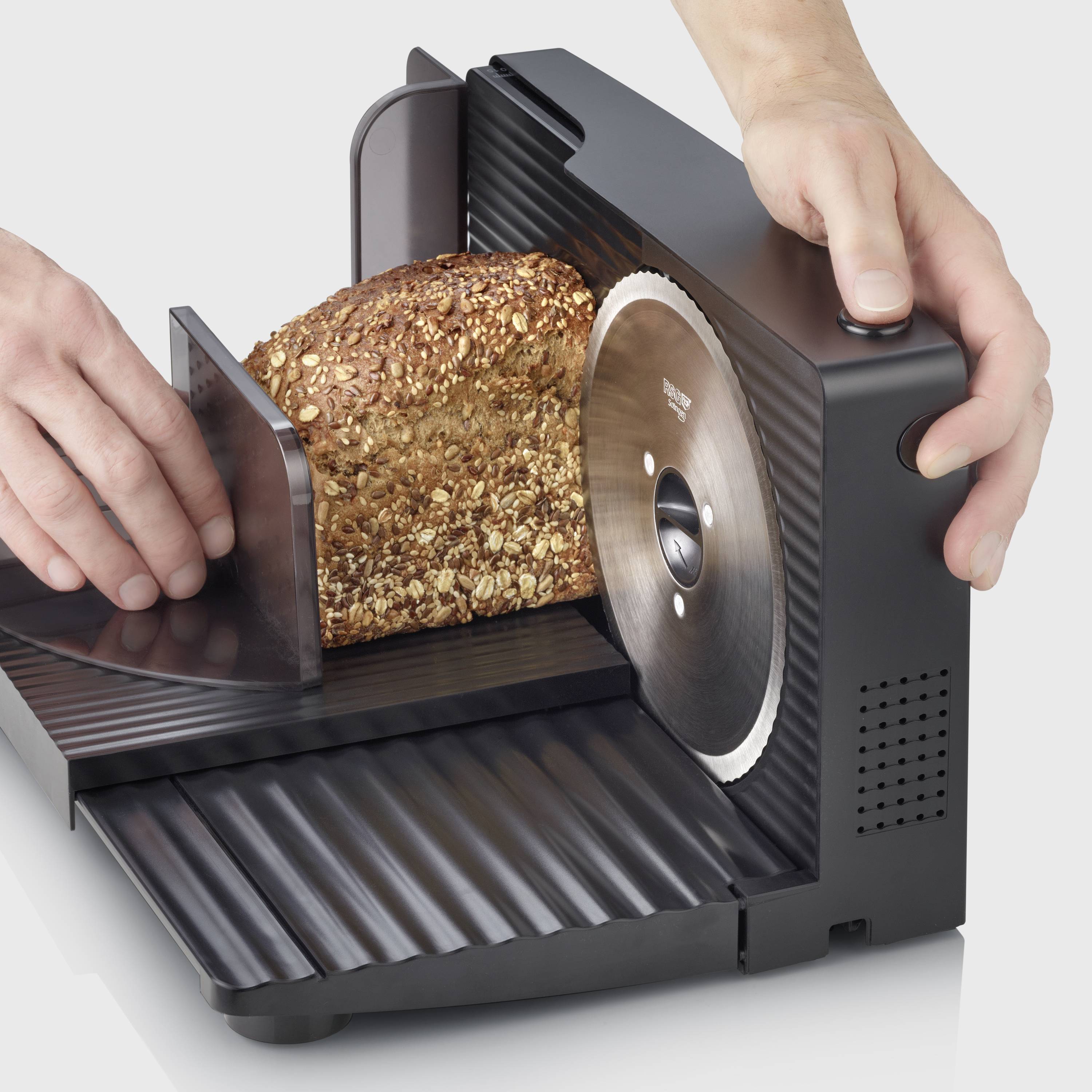 Hleb polnozrnatega kruha se reže z električno napravo za rezanje kruha. Ena roka vodi kruh proti rezilu.