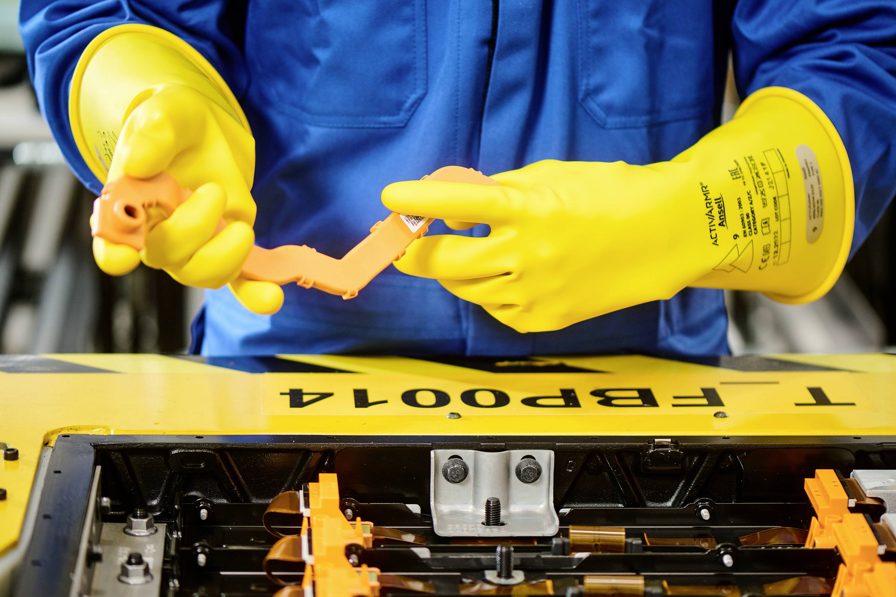 Oseba v modri delovni obleki in rumenih zaščitnih rokavicah dela na odprtem prostoru za baterijo z električnimi orodji.