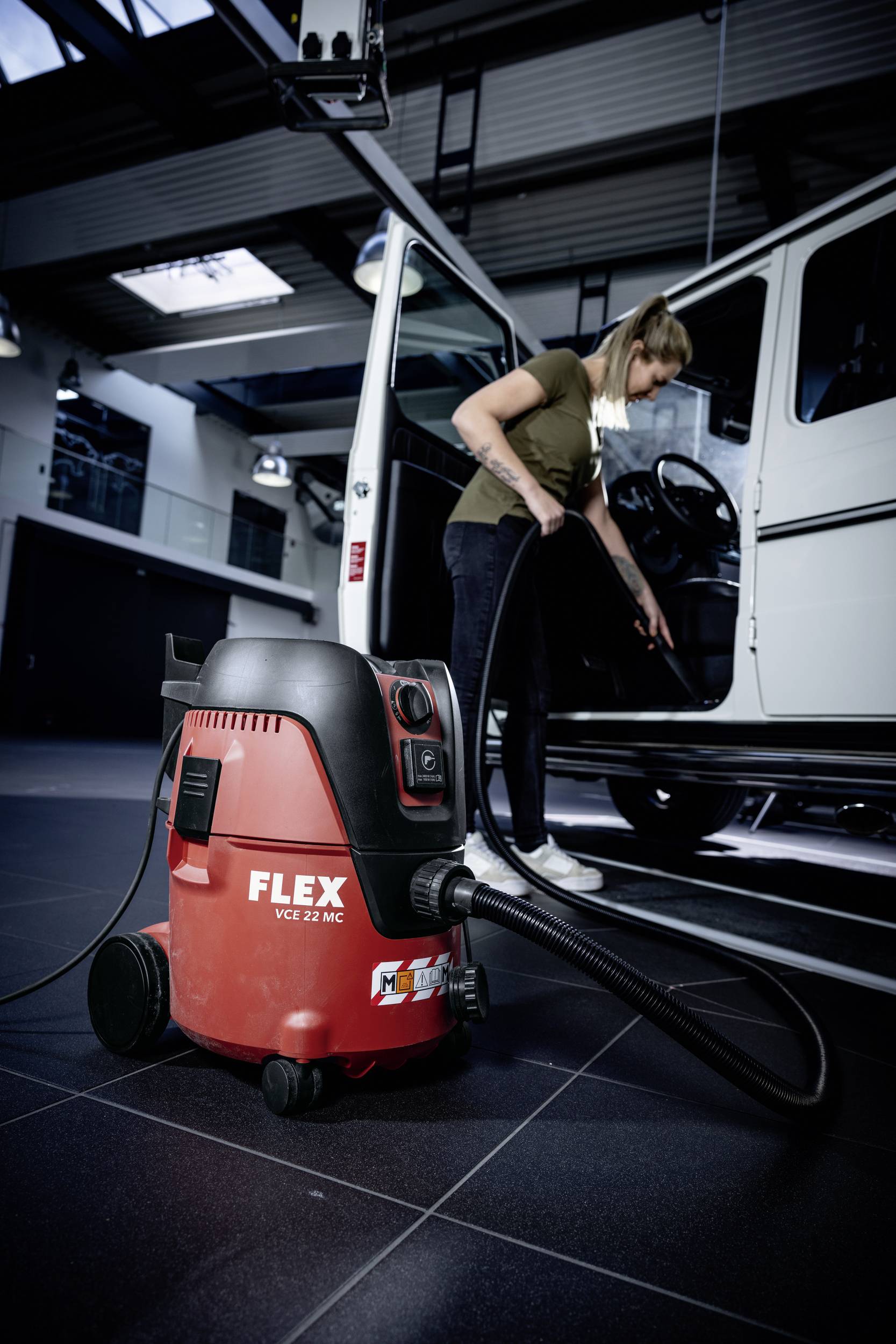 Oseba čisti notranjost avtomobila z rdečim sesalnikom znamke Flex. Prostor deluje kot avtomehanična delavnica.