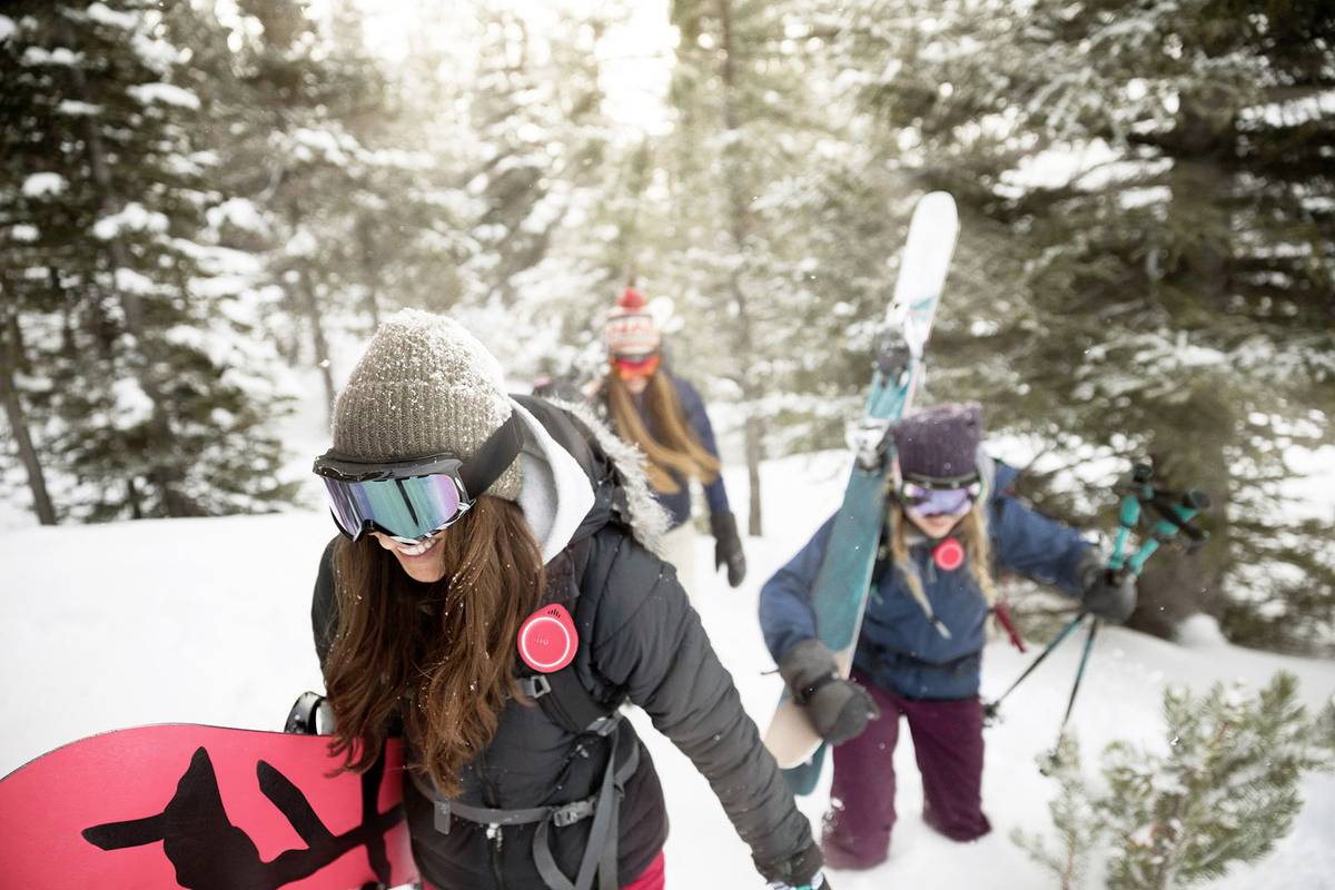 Skupina treh ljudi se sprehaja skozi zasnežen gozd, s seboj nosi opremo za zimske športe, vključno s snowboardom in smučmi. Oblečeni so v topla oblačila.