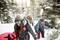 Skupina treh ljudi se sprehaja skozi zasnežen gozd, s seboj nosi opremo za zimske športe, vključno s snowboardom in smučmi. Oblečeni so v topla oblačila.