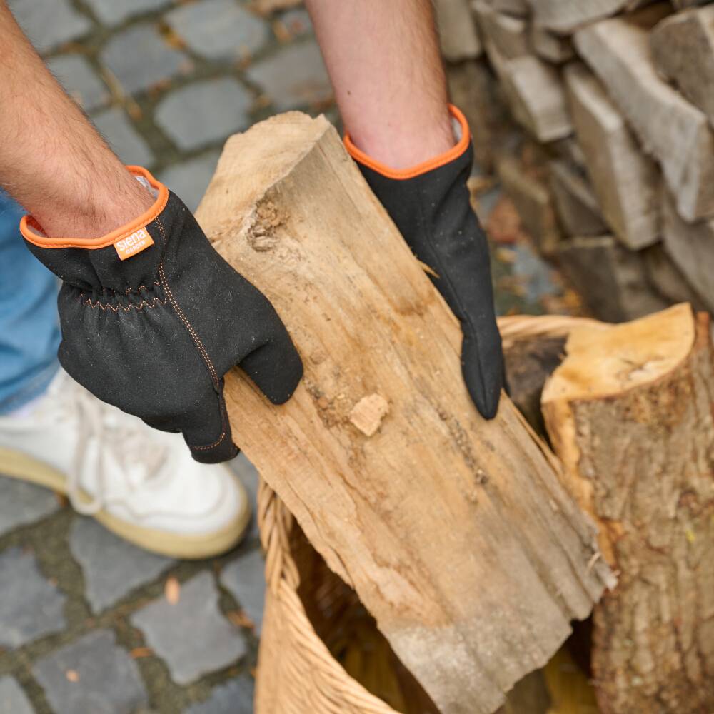 Oseba dviguje hlodovino iz pletene košare. Oblečene ima črne rokavice z oranžnimi šivi.