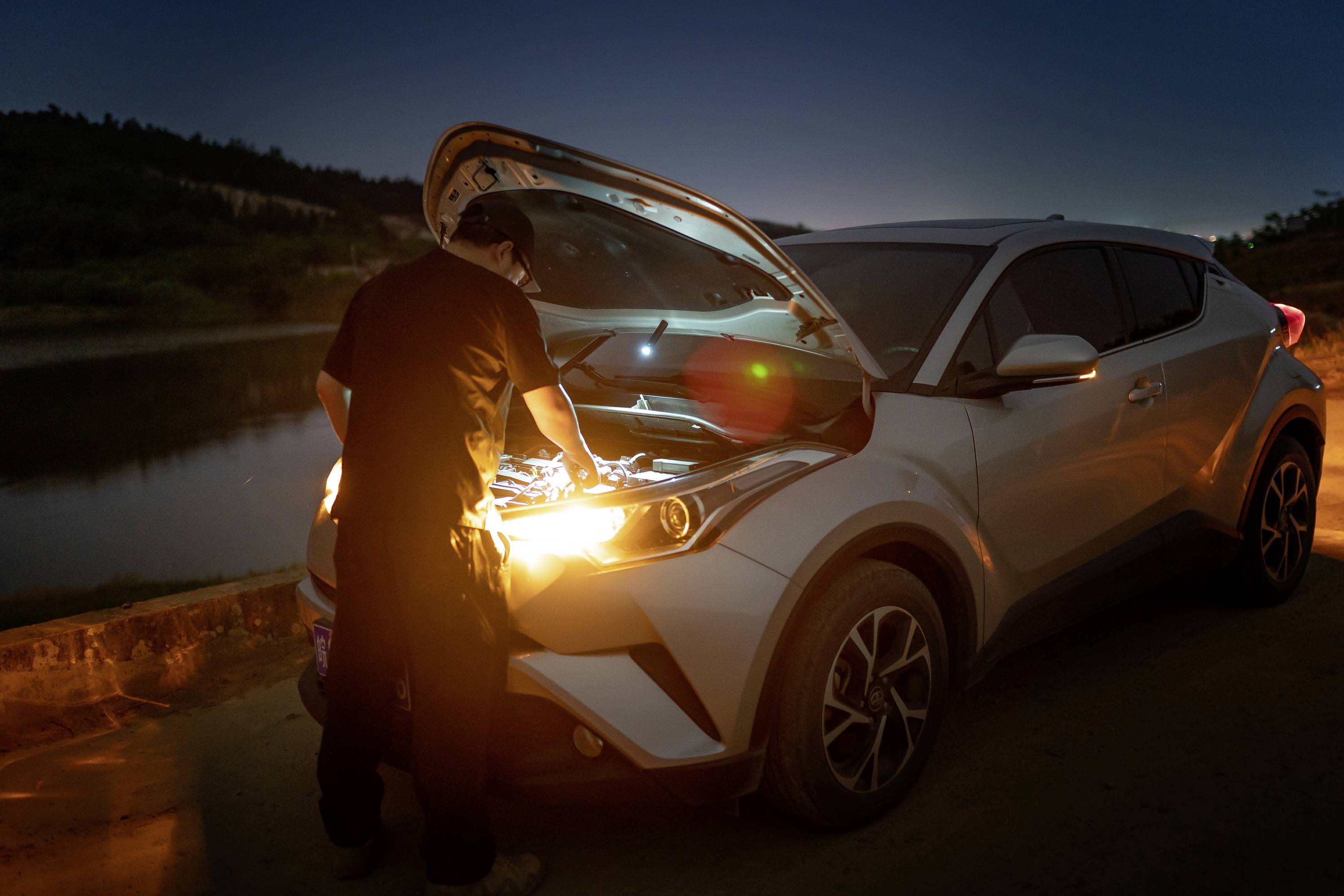 Oseba popravlja avto ponoči ob cestišču, odprta je maska motorja, svetloba posvetljuje motor.