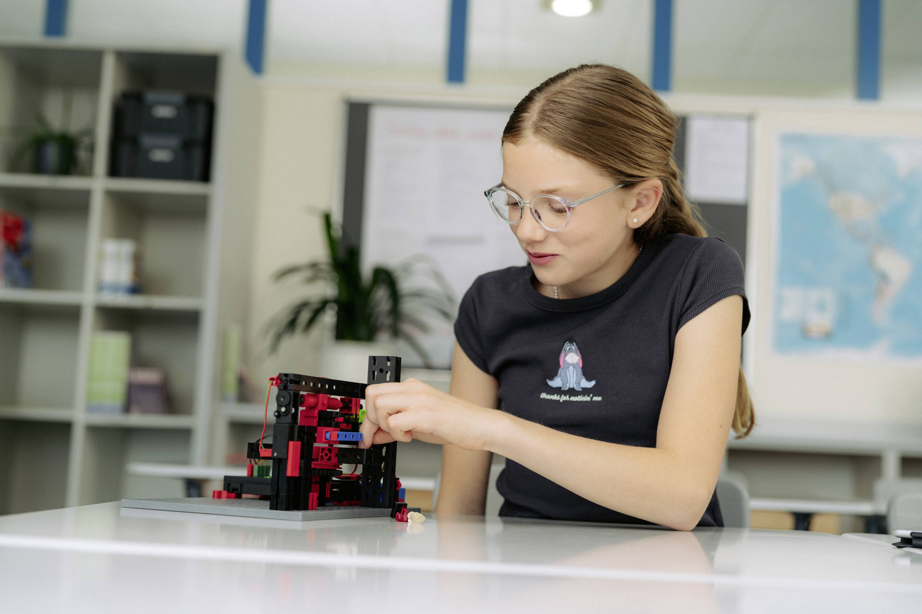 Dekle z očali gradi LEGO robota v učilnici. V ozadju so police in svetovna karta.