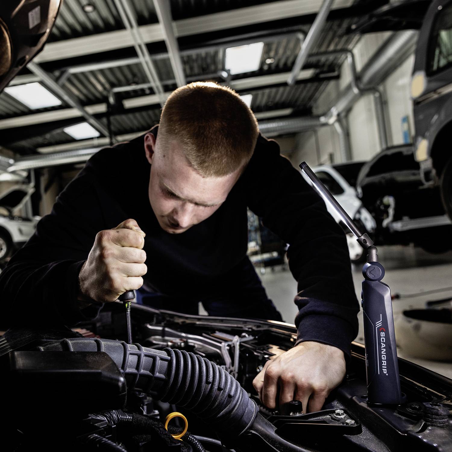 Mehanik s kratko pričesko popravlja motor v delavnici, osredotočen na delo, z moderno opremo v ozadju.