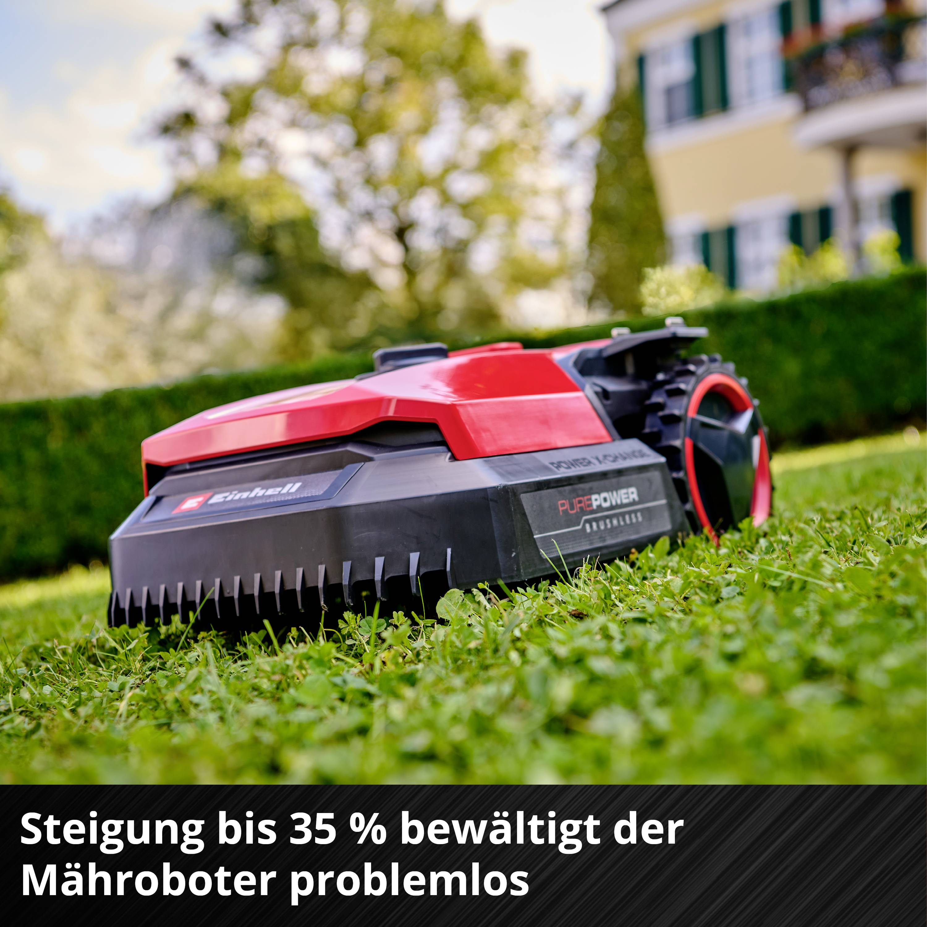 Robotska kosilnica kosi travo pred hišo. Besedilo: "Robotska kosilnica brez težav premaga naklone do 35 %."