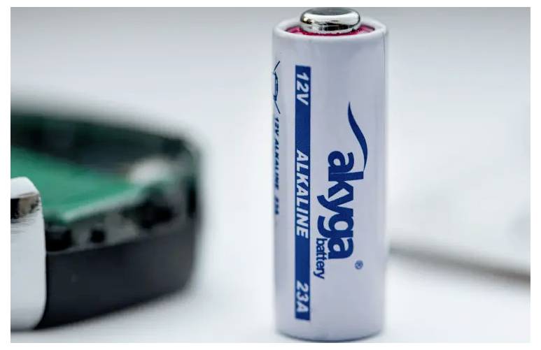 Bela, cilindična 12V alkalna baterija z napisom 'akyga battery' stoji poleg delno vidne zelene naprave.