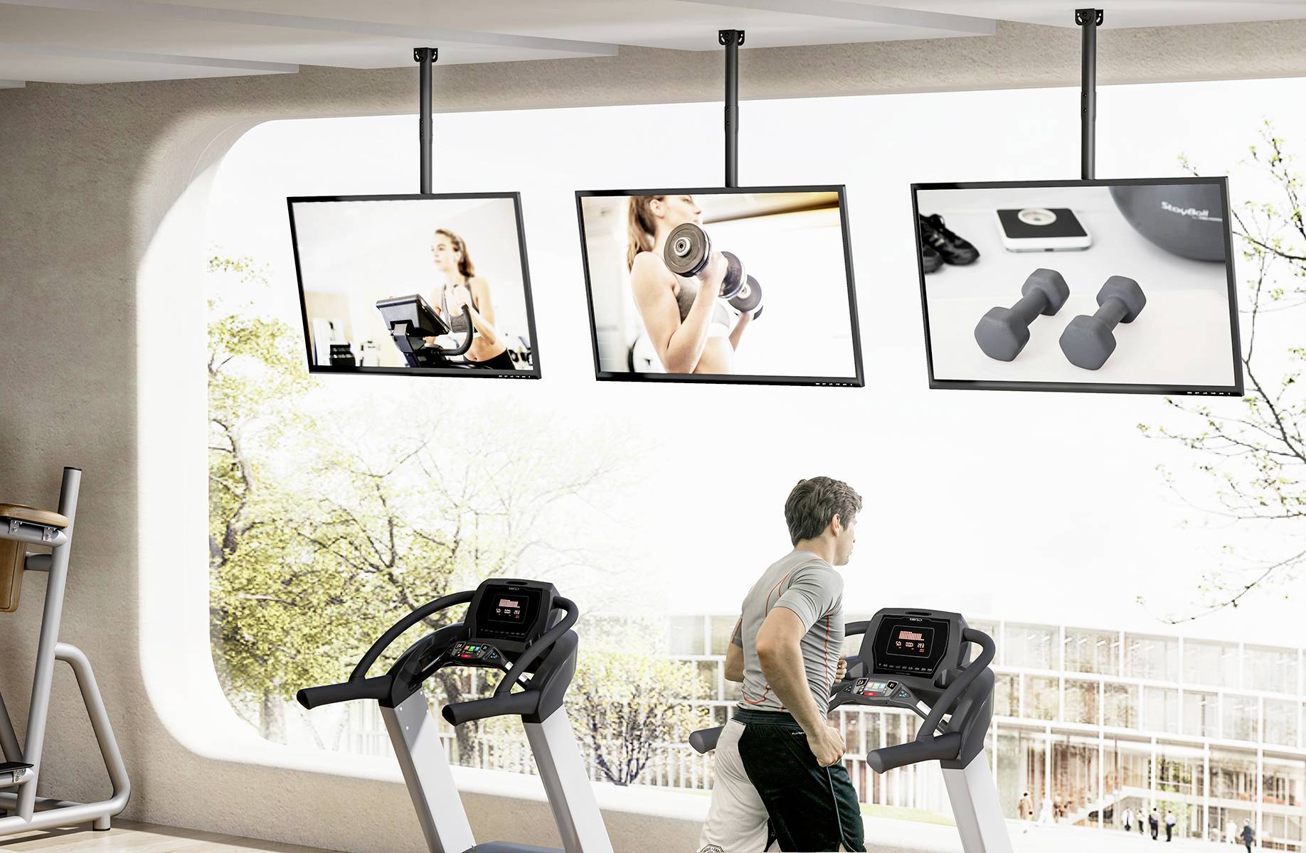 Moški teče na tekalni stezi v fitnesu. V ozadju so trije veliki zasloni s fitnes podobami.