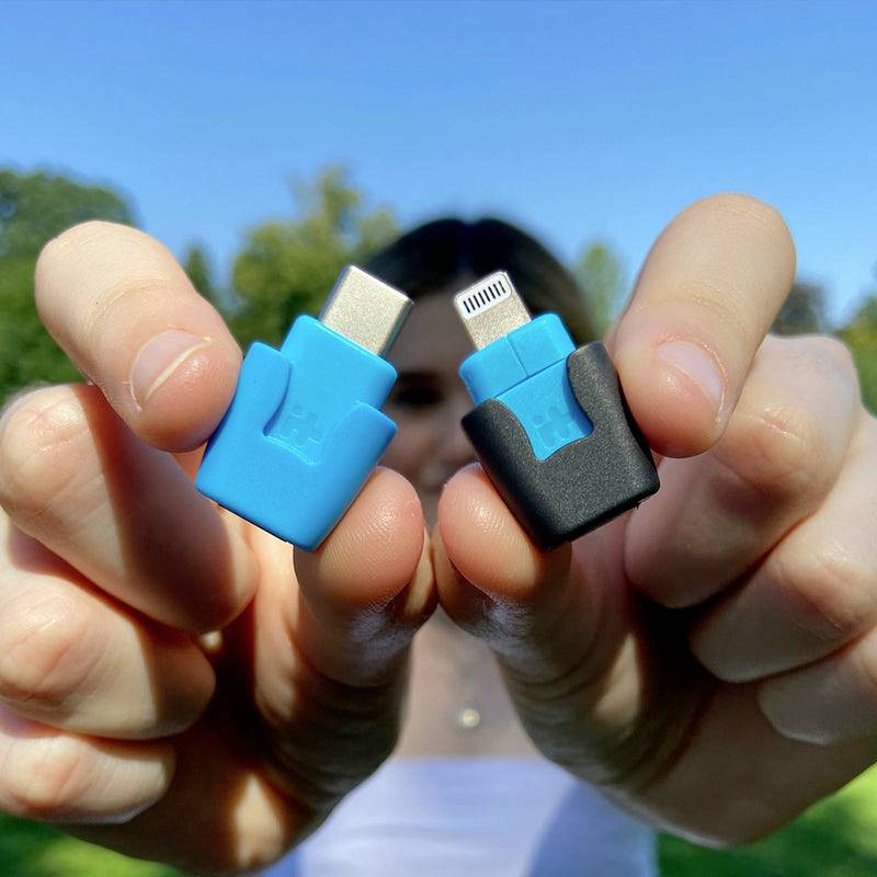 Oseba drži dva adapterja, enega modrega USB-C adapterja in enega črnega Lightning adapterja, pred zamegljenim ozadjem.