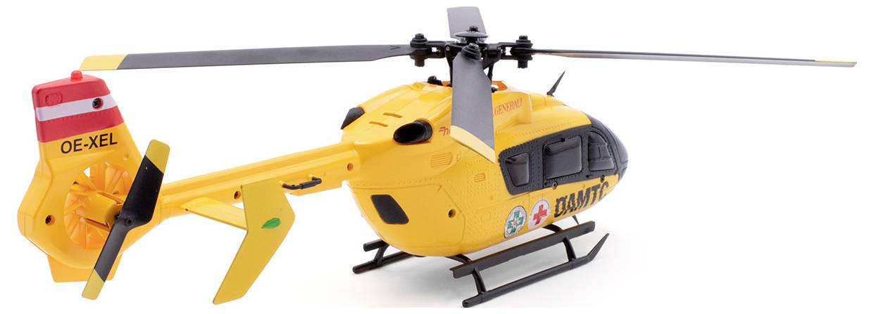 Rumen modelni helikopter z rdečimi in črnimi poudarki stoji na tleh, z vidnimi nalepkami in oznakami.