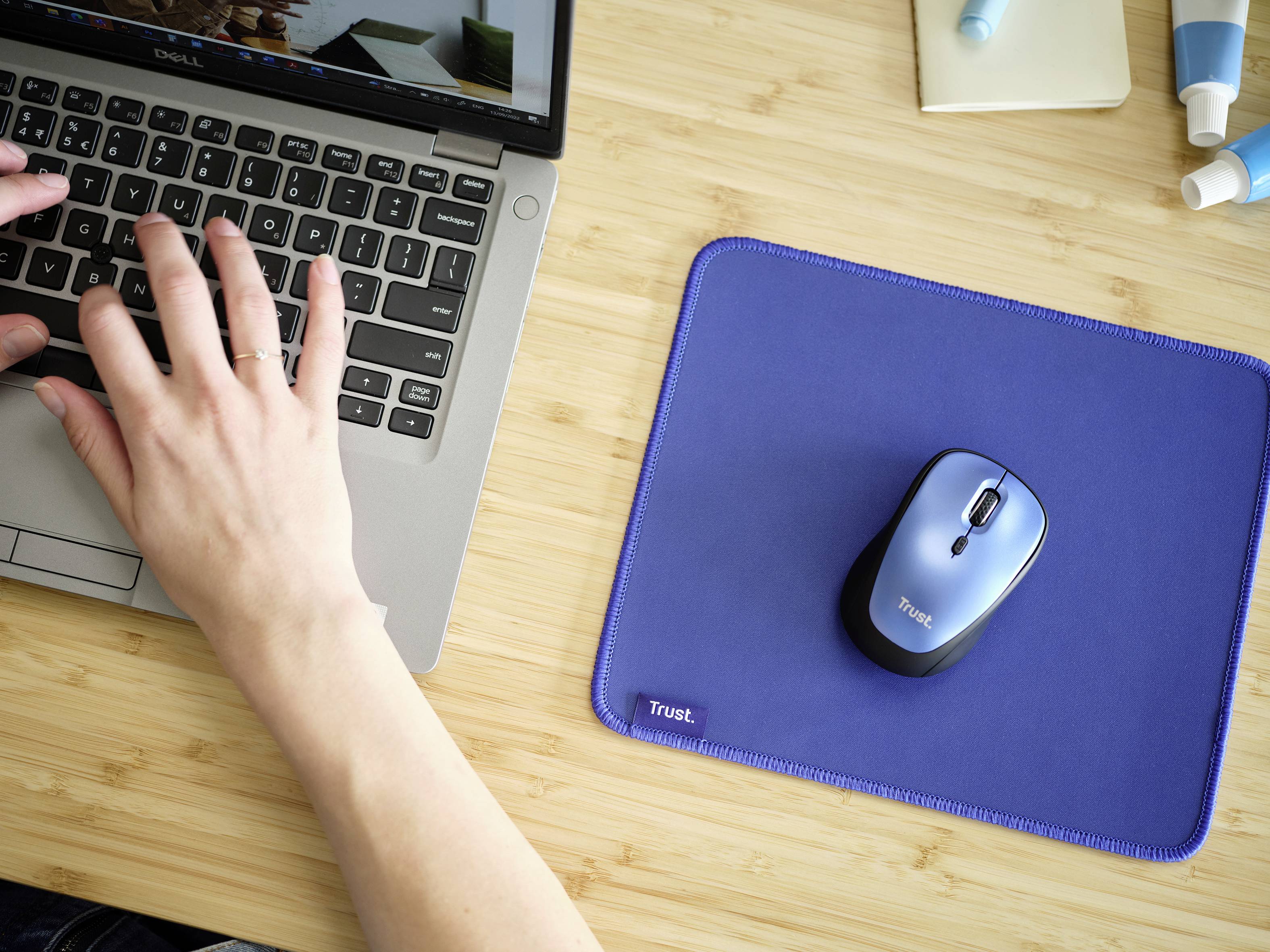 Roke tipkajo po tipkovnici prenosnika. Zraven leži modra podloga za miško z modro miško na leseni mizi.