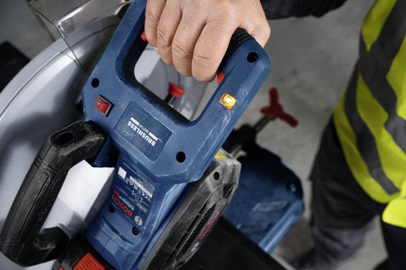 Oseba drži električno orodje z modrim ohišjem, verjetno žago, v desni roki. Nosi zaščitno opremo.