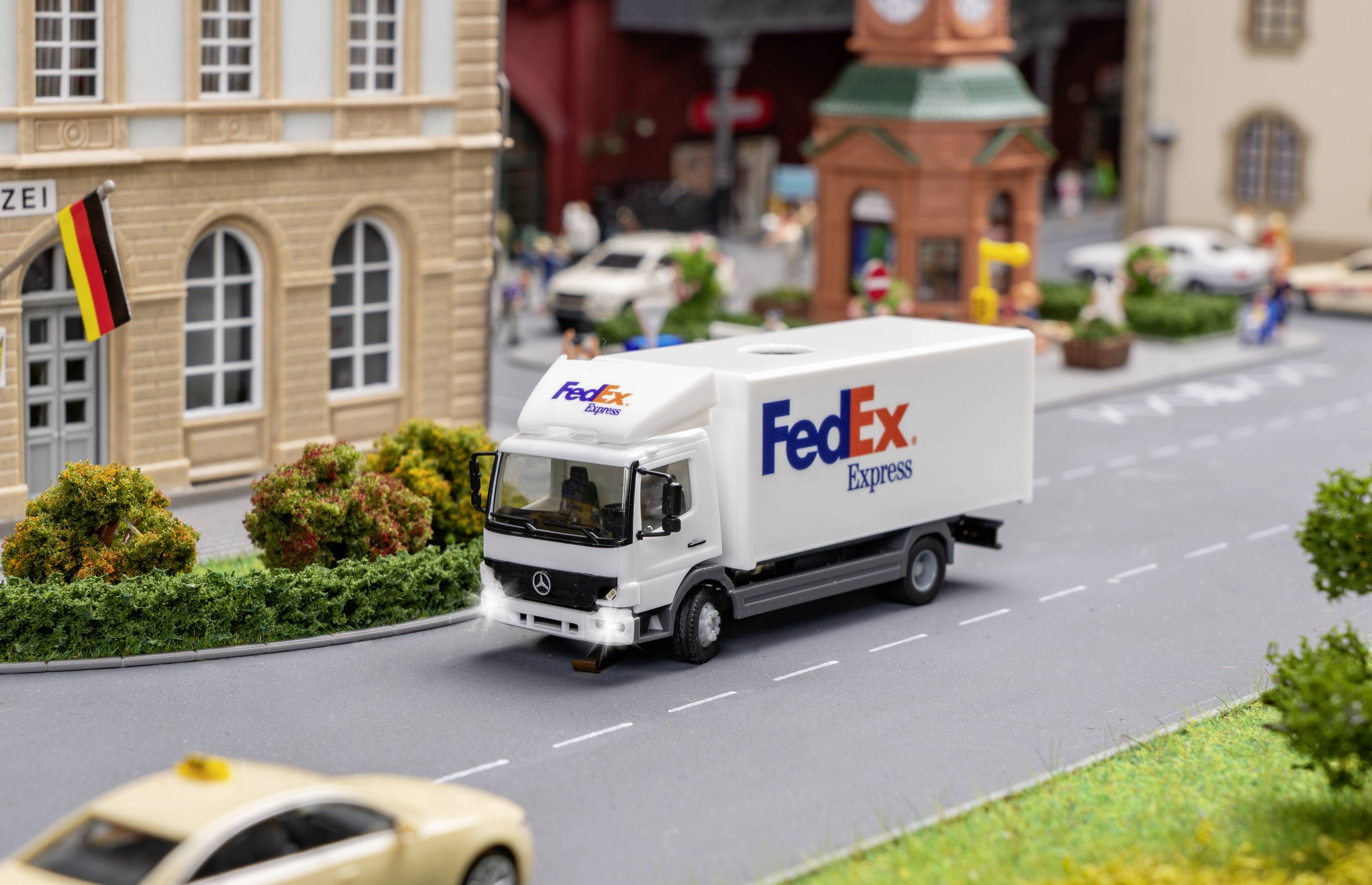 Bel dostavni kombi podjetja FedEx Express vozi skozi miniaturni model mesta z avtomobili in stavbami v ozadju.